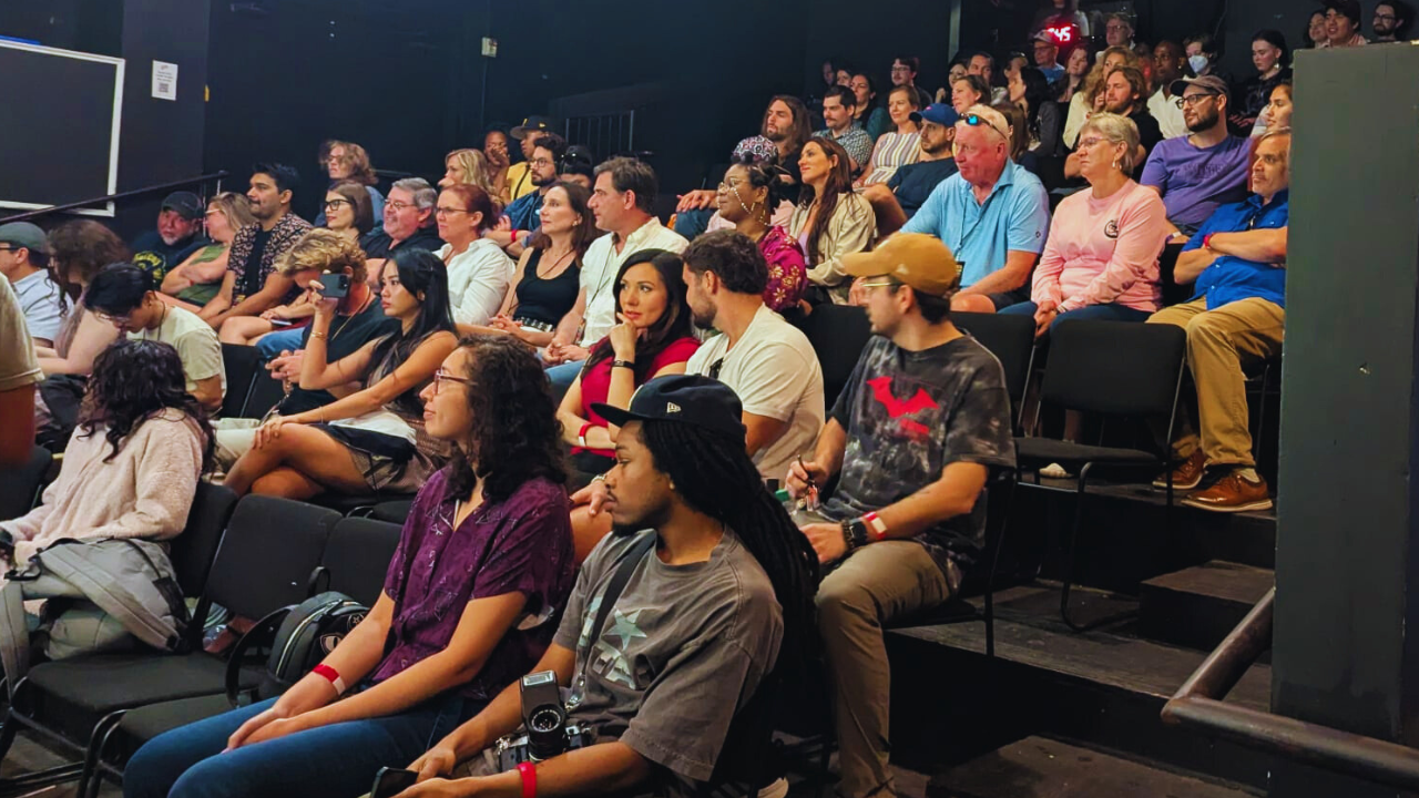Audience seated in a dark theater or auditorium, attentively watching a presentation or performance.