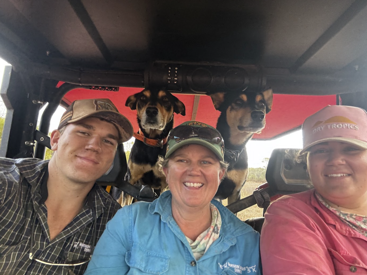 Three people and two dogs inside a vehicle, with two on the left and one on the right, smiling, two wearing hats, and the dogs in the background.