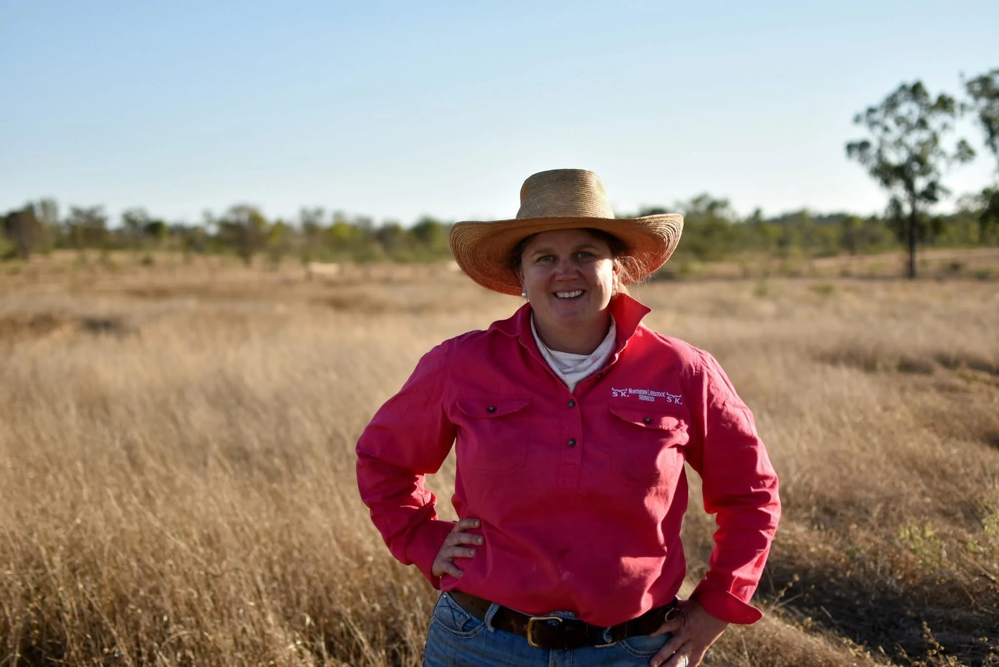 A woman wears a wide-brimmed hat, pink shirt, and blue jeans, standing in a dry field with scattered trees under a clear sky.