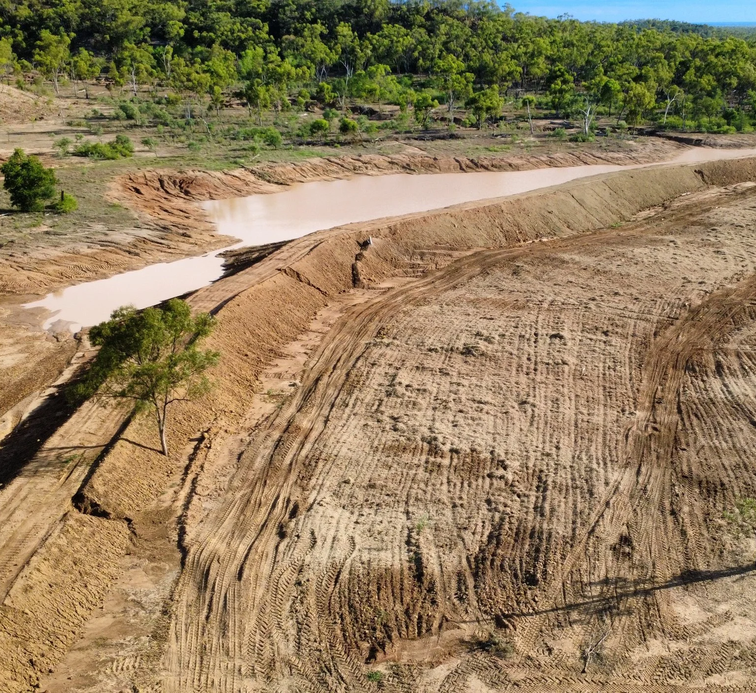 Aerial view of a construction site with uneven dirt and soil, a small tree, and a body of water in the background surrounded by green trees.