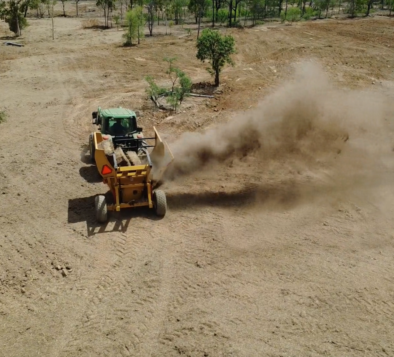 A green tractor pulling a large yellow soil grader dispersing dirt on a dry, cleared landscape with some scattered trees.