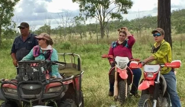 Four people outdoors with motorcycles and a utility vehicle in a grassy field with trees and cloudy sky in the background.