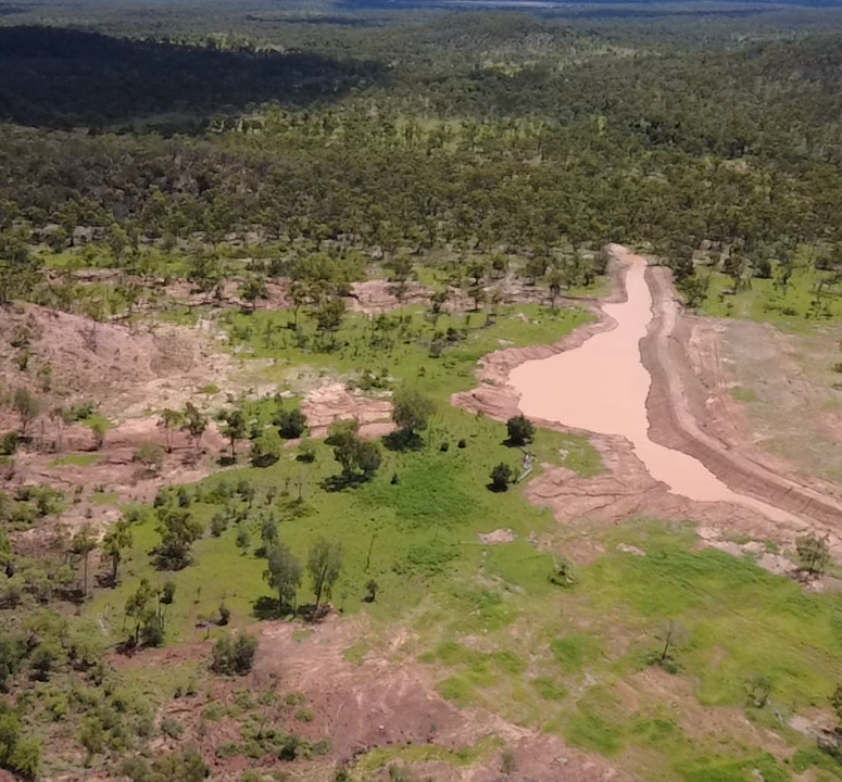 Aerial view of a landscape showing a muddy river flowing through green and pinkish eroded land, with patches of trees and shrubs, and forested horizon in the background.