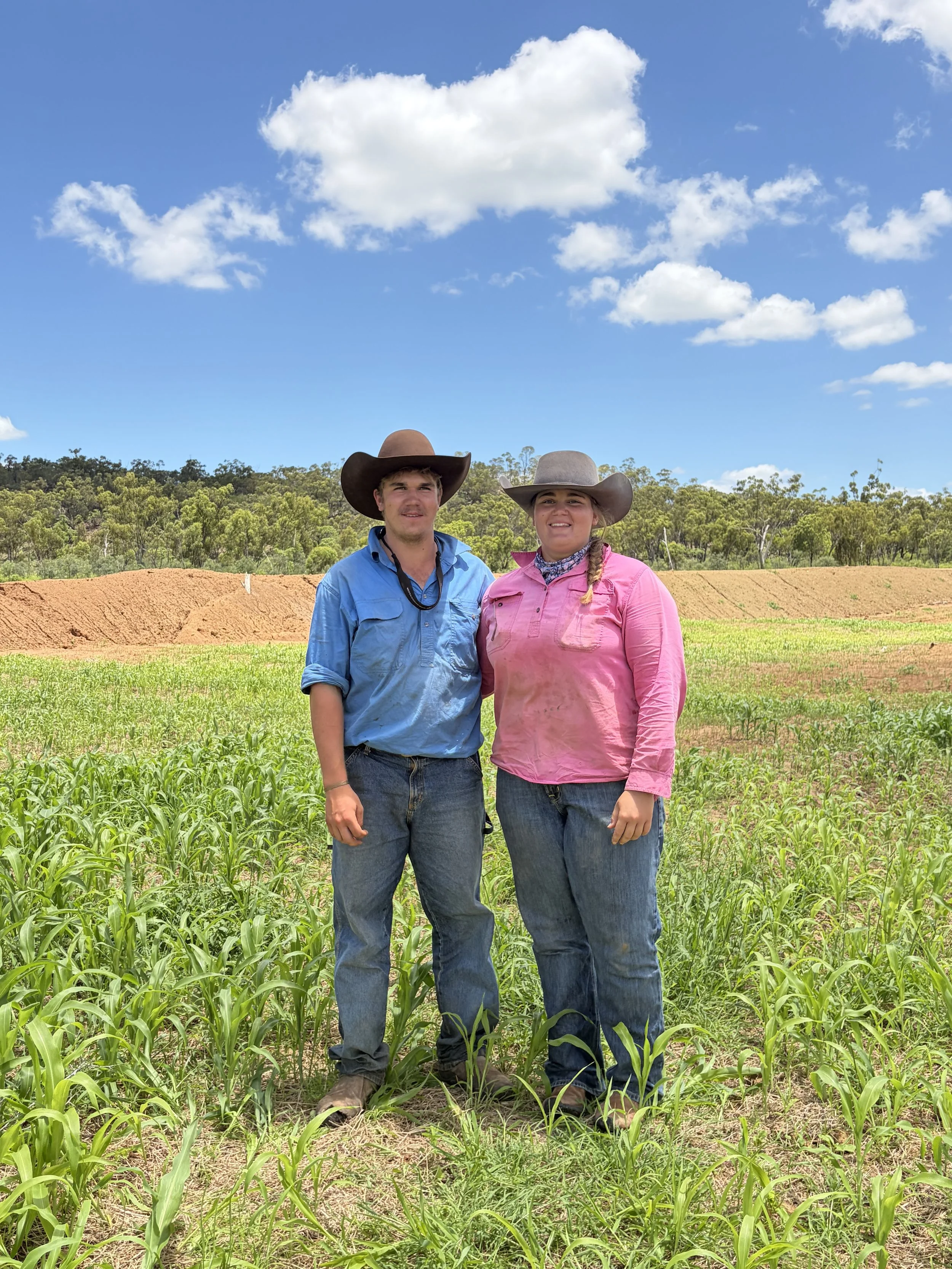 Two farmers, a man and a woman, standing side by side in a green field under a bright blue sky with scattered clouds. Both are wearing wide-brimmed hats, casual shirts, and jeans, smiling at the camera.