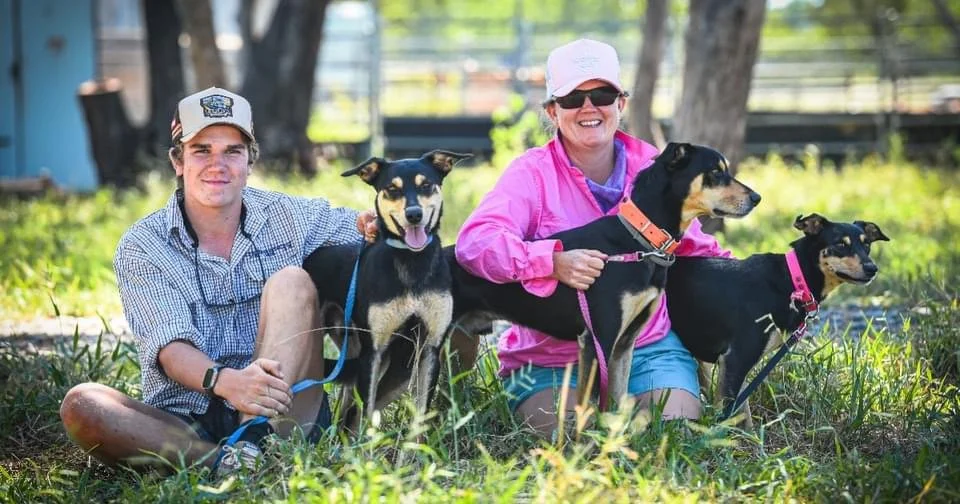 A young man and an older woman sitting outdoors with three black and tan dogs, all on leashes, in a grassy area with trees in the background.