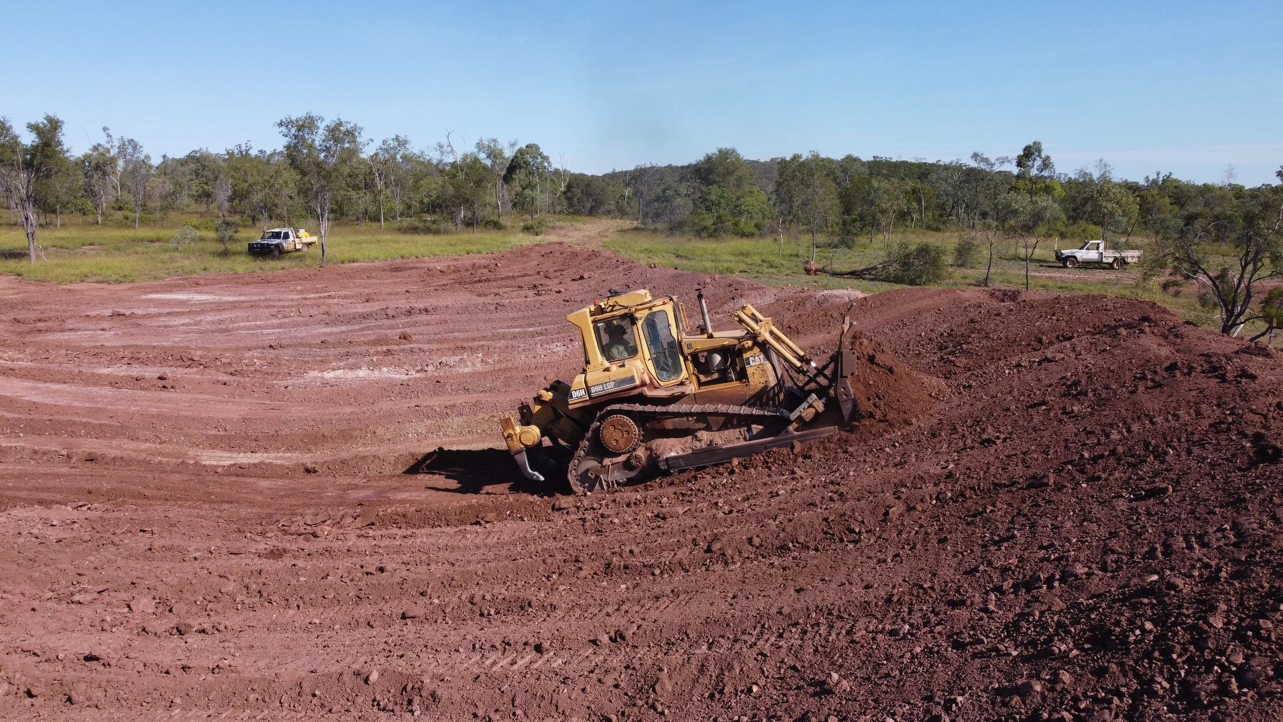 A bulldozer is leveling the ground on a construction site, with trees and two pickup trucks in the background under a clear blue sky.