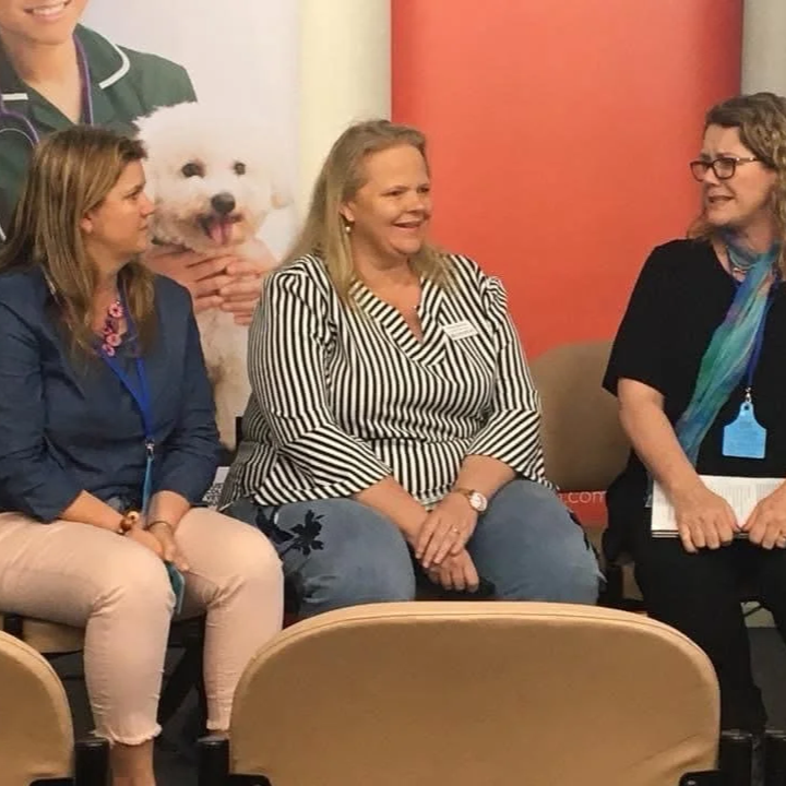 Three women sitting together at an event, with a large poster of a fluffy white dog in the background.