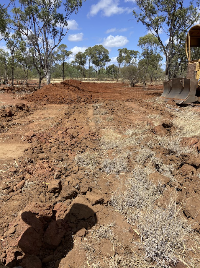 A construction site in a dry, rural area with freshly dug dirt and sparse dry vegetation. There is a bulldozer on the right side and trees in the background under a partly cloudy sky.
