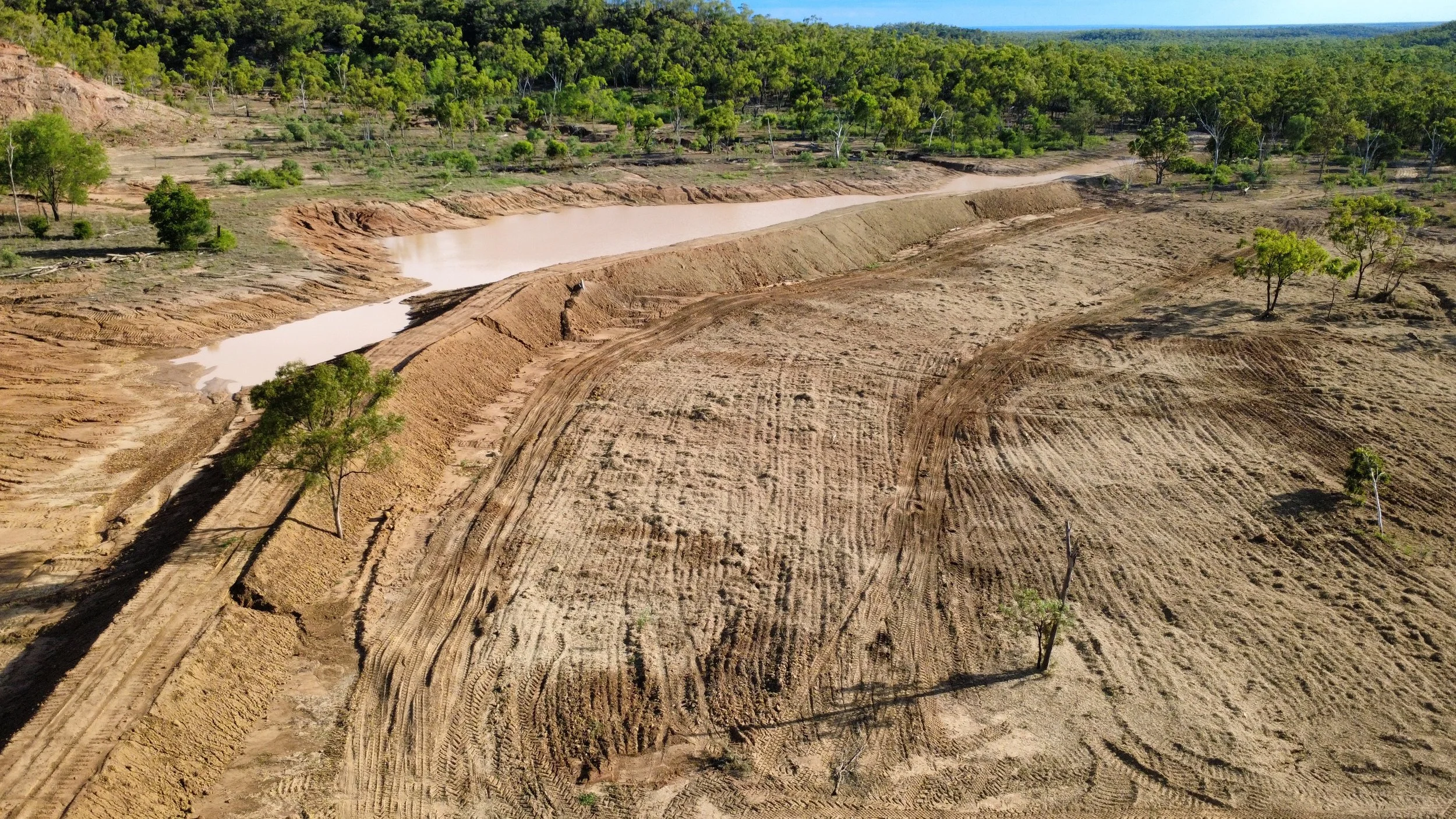 An aerial view of a construction site with earthwork, a water-filled trench, and sparse trees, with a wooded area in the background.