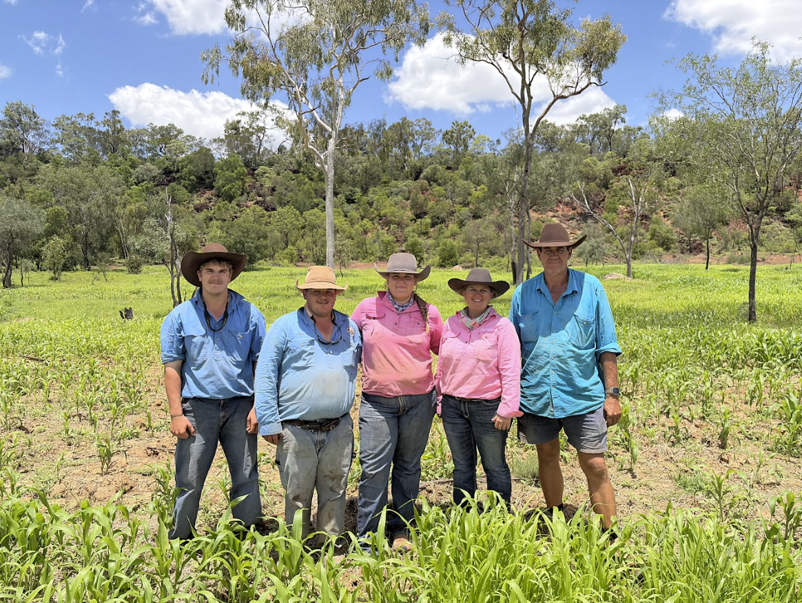 Five people standing in a field of young crops, wearing wide-brimmed hats, long sleeves, and jeans, with a background of trees, hills, and a partly cloudy sky.