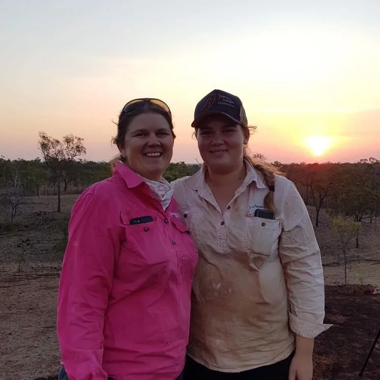 Two women standing outdoors at sunset, smiling, with a landscape of trees and a setting sun in the background.