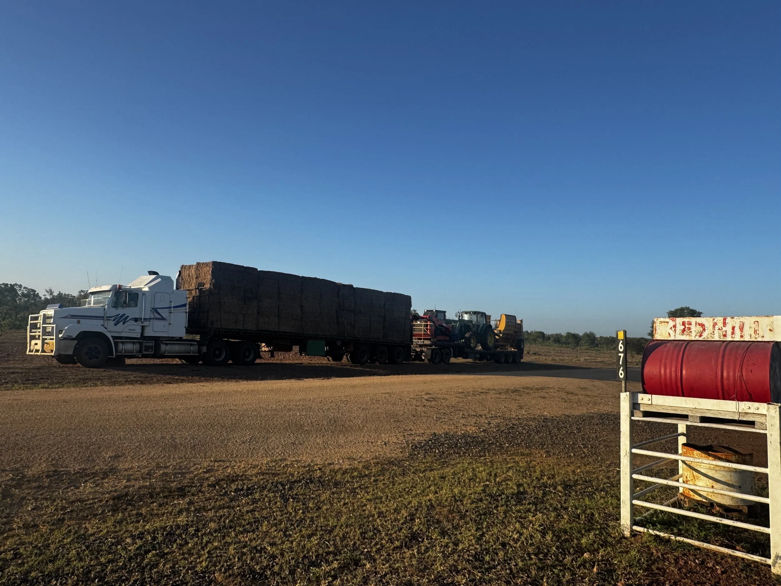 A white semi-truck carrying a load of hay bales on a rural dirt road with a tractor and farm equipment in the background, under a clear blue sky.