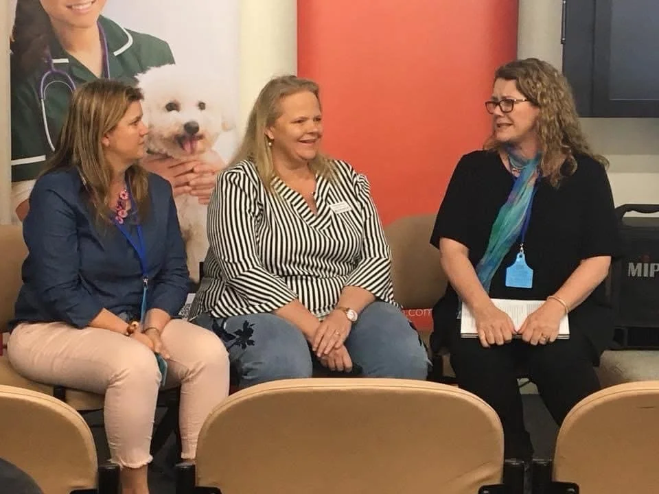 Three women sitting on a beige couch, engaged in conversation. There is a large poster of a white dog with a woman holding it, visible in the background.