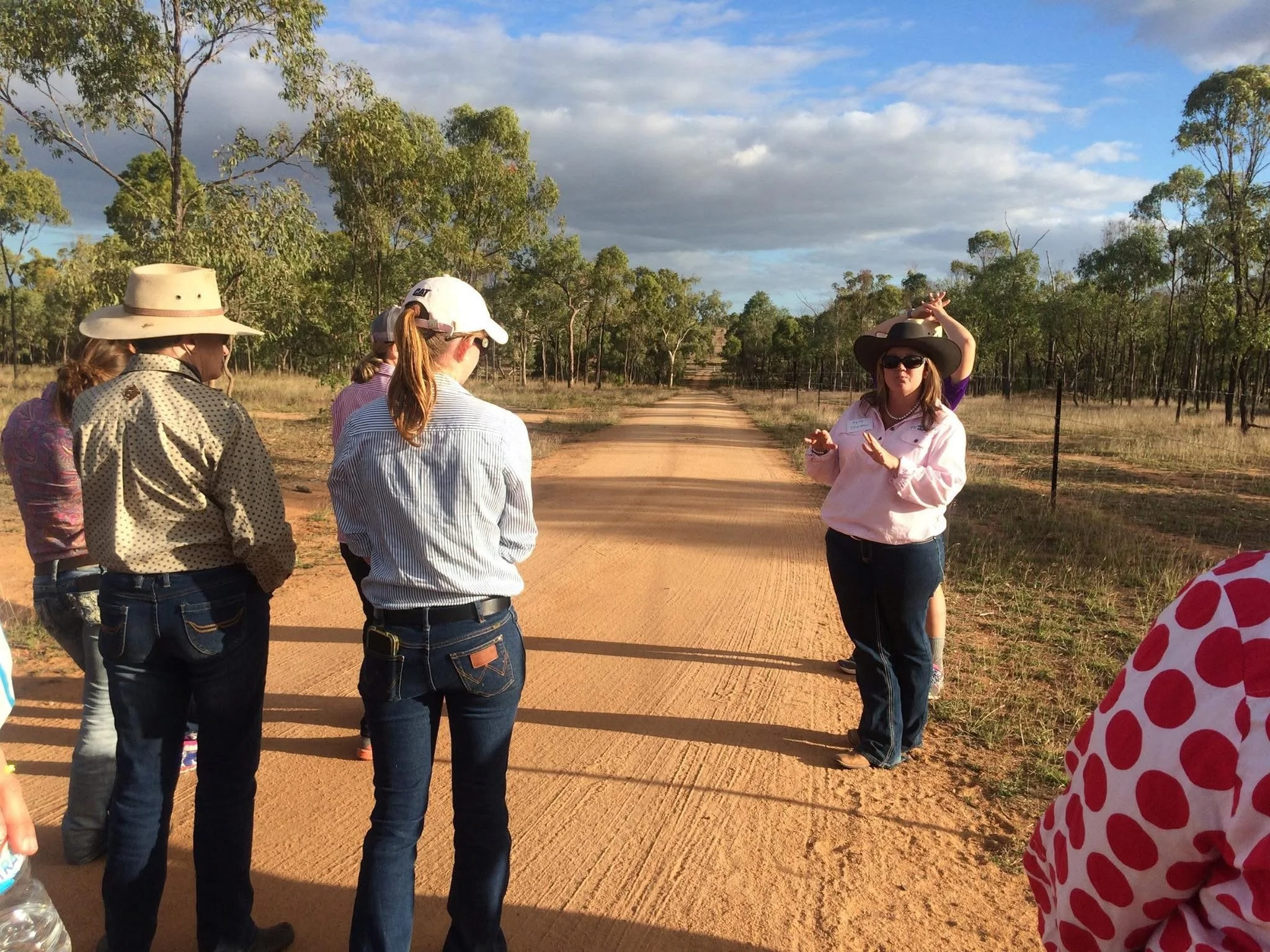 A group of people standing on a dirt road in a rural area with trees on either side, listening to a woman wearing a wide-brimmed hat and sunglasses, who appears to be giving a tour or lecture.