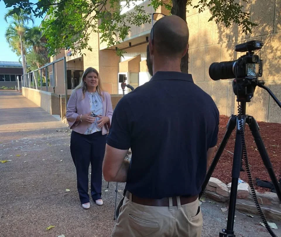 A woman being interviewed outdoors by a man with a camera on a tripod, with a building and trees in the background.