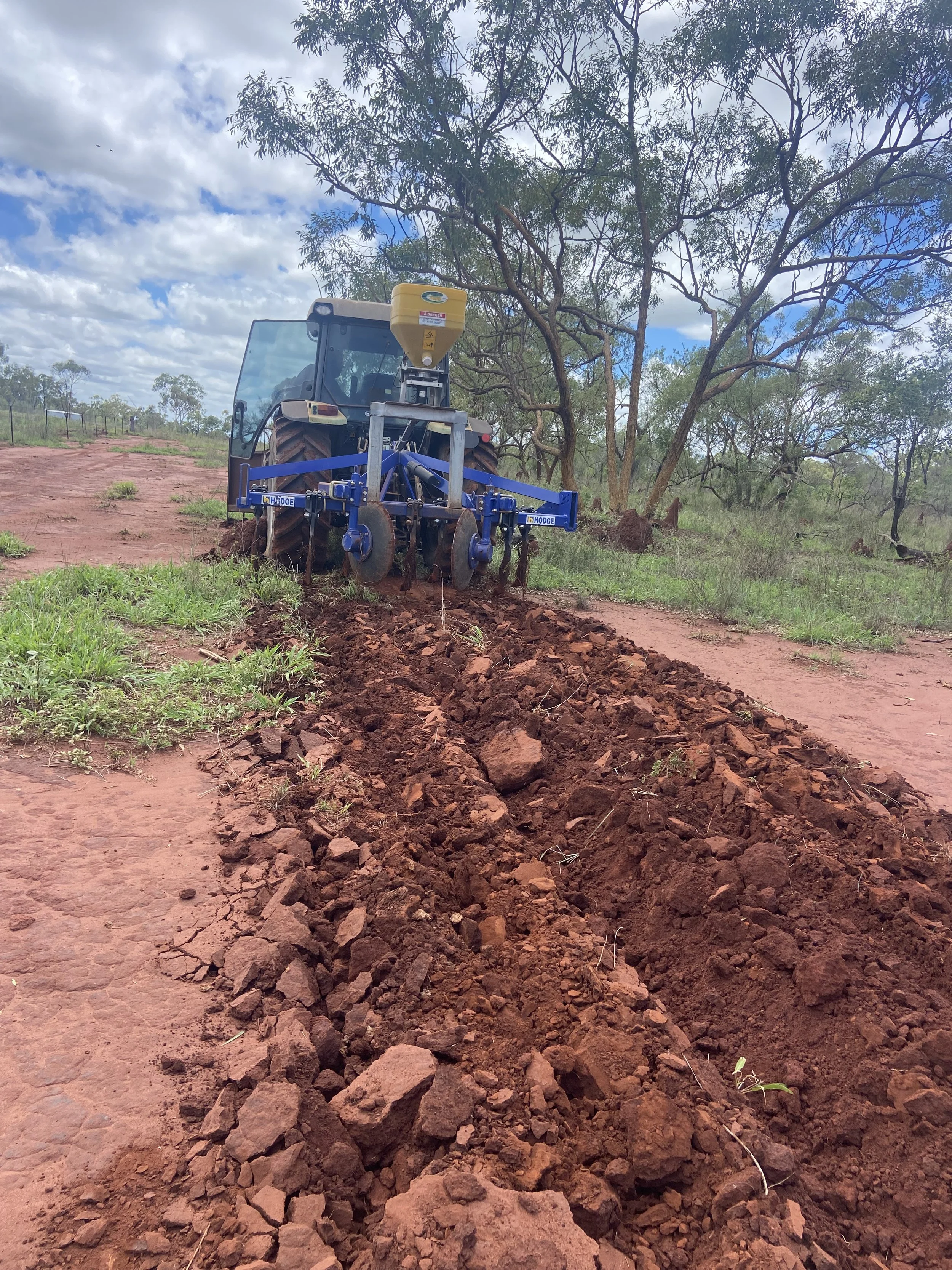A tractor working on red soil in a rural area, with green trees and a partly cloudy sky in the background.