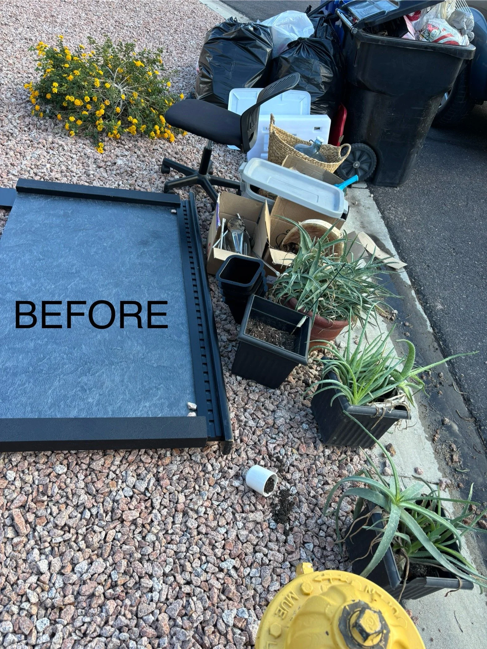 An area with a large black trash bin, filled with bags of trash, and a yellow fire hydrant. Outside, there are scattered plants, boxes, and gardening supplies, along with a black office chair missing its base, on a gravel surface next to a curb.