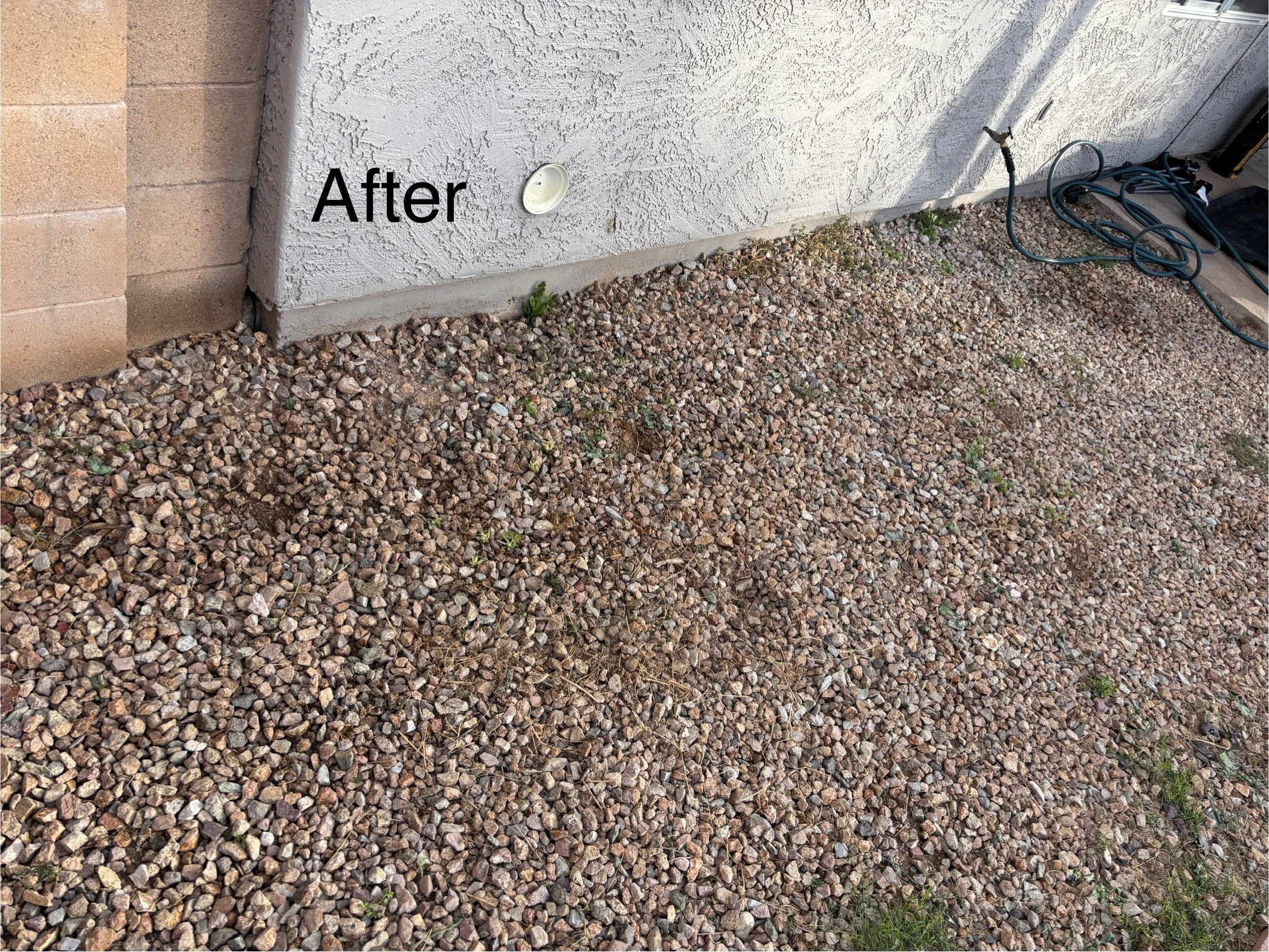 Side of a house with a textured white wall, a small round light fixture, and a sprinkler hose on gravel ground. The word 'After' is visible on the wall.