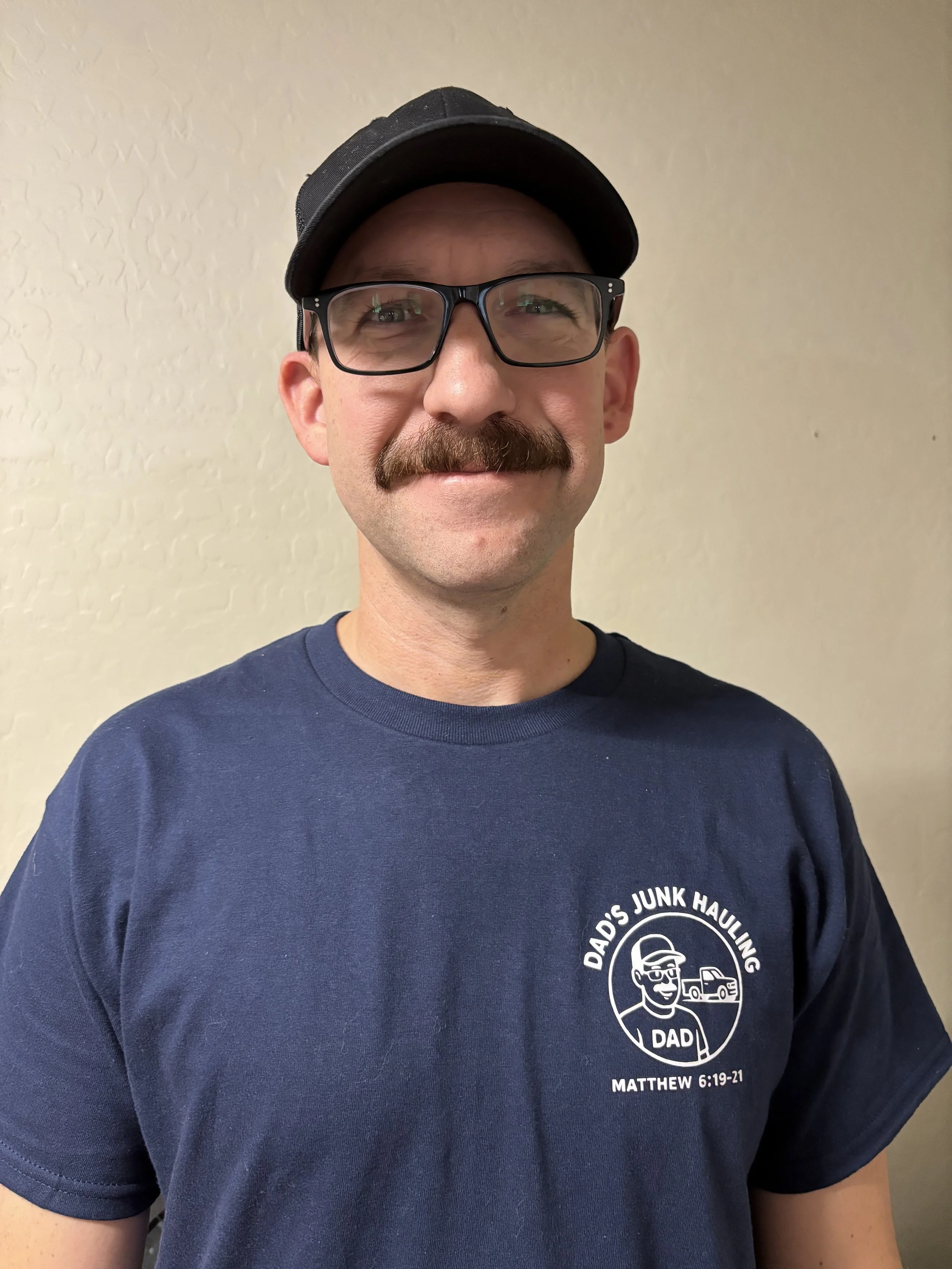 A man with glasses, a mustache, wearing a cap and a navy blue T-shirt that reads 'Dad's Junk Hauling' with a graphic of a man and vehicles, standing against a beige wall.