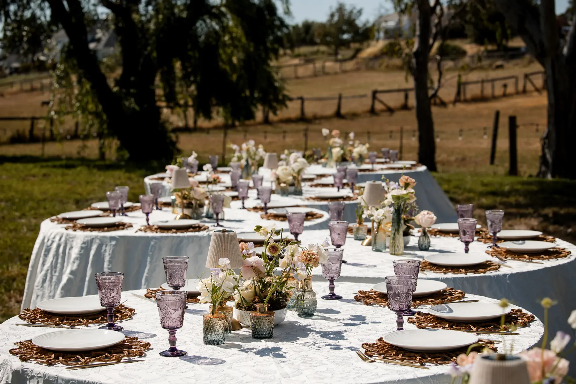 Outdoor dining tables set with white tablecloths, floral centerpieces, purple glassware, and white plates with woven placemats, set in a grassy area with trees in the background on a sunny day.