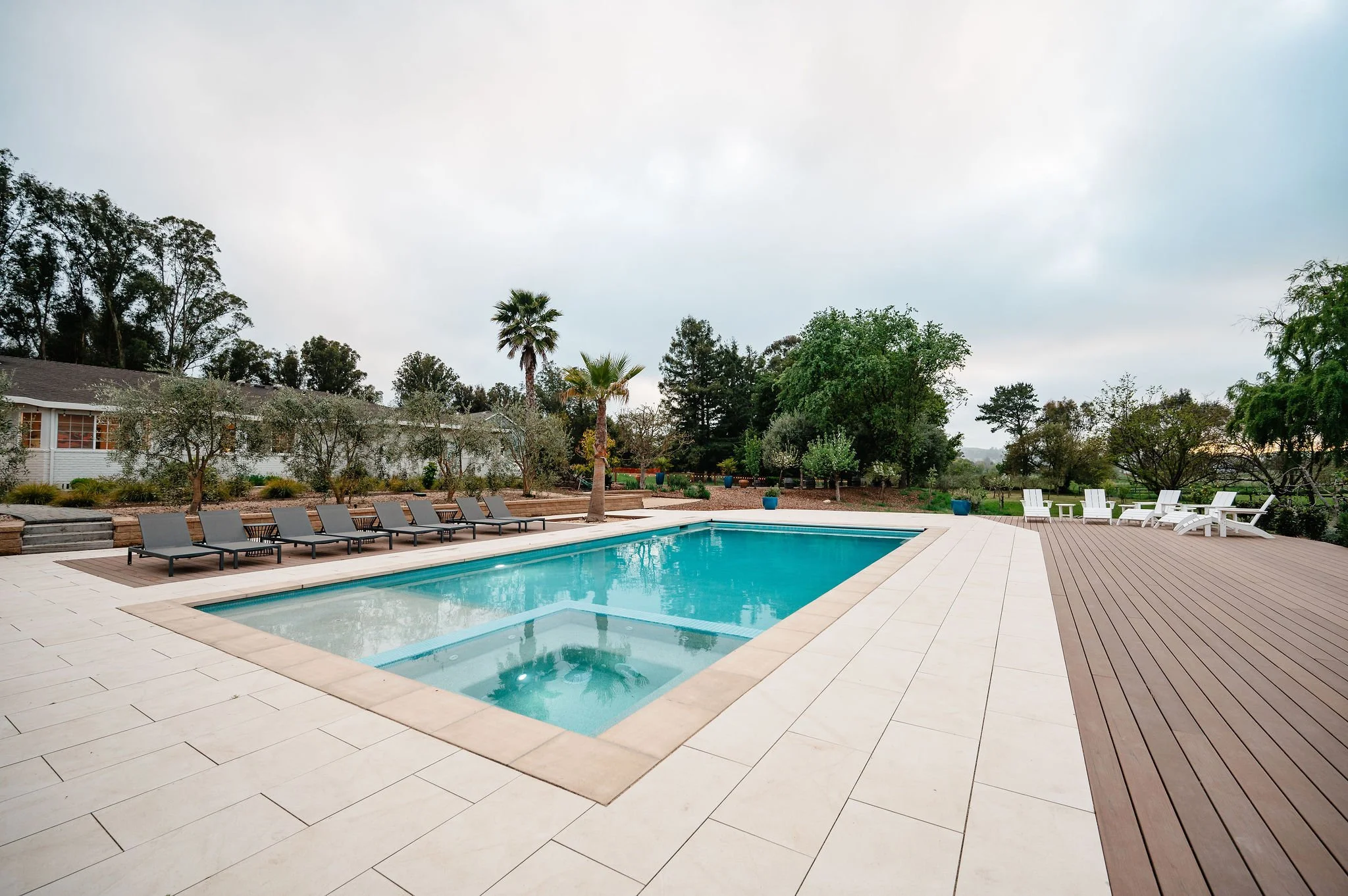 Empty swimming pool with a surrounding deck area, lounge chairs, and trees in the background on a cloudy day.