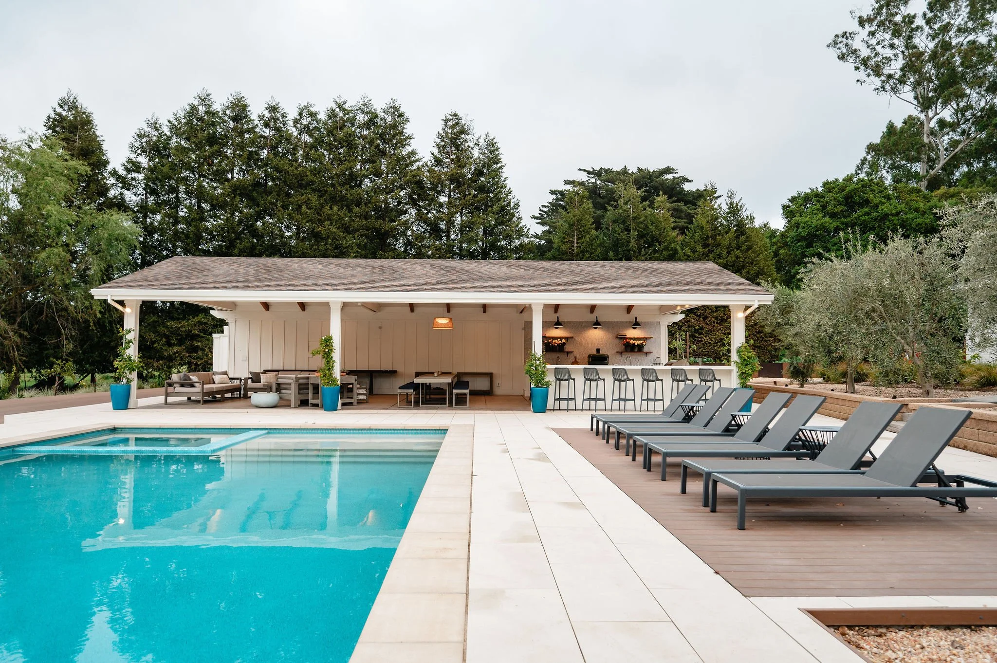 Empty outdoor pool area with lounge chairs, a bar, and seating area, surrounded by trees and landscaping.