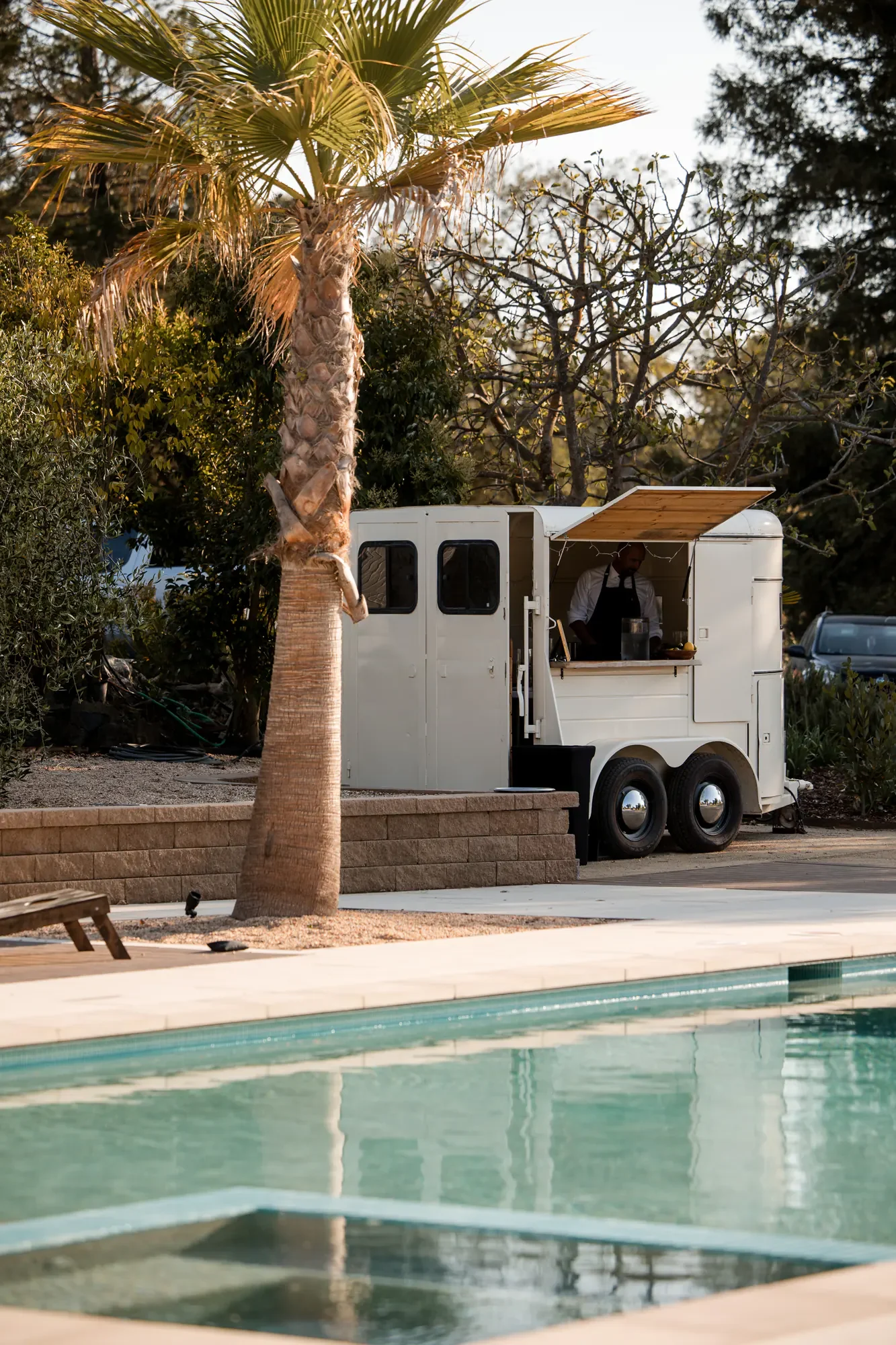 A white food truck with an awning, parked near a pool and a palm tree, with a man inside preparing food.