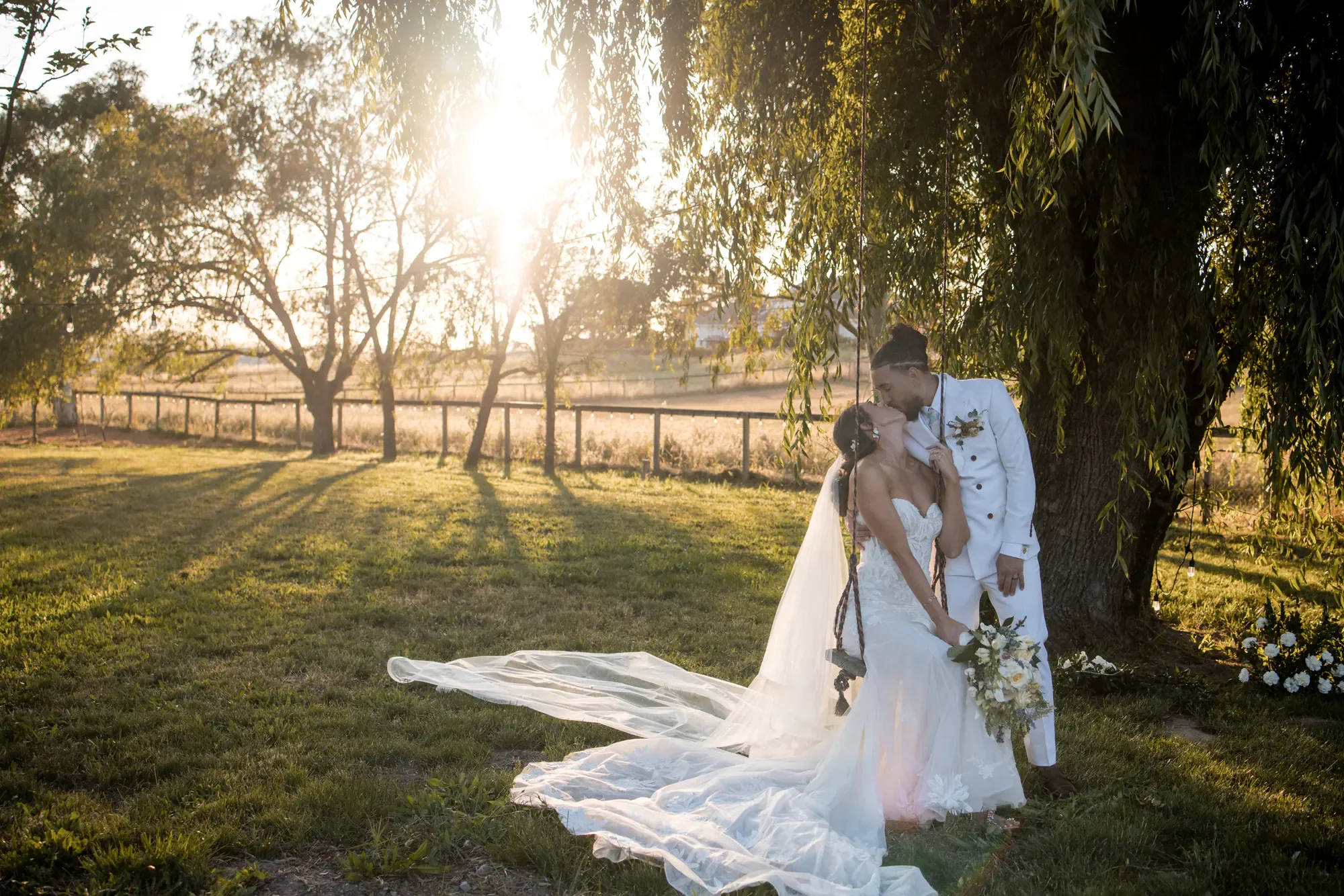 A bride and groom sharing a kiss outdoors under a large tree with sunlight shining through, during their wedding photoshoot.