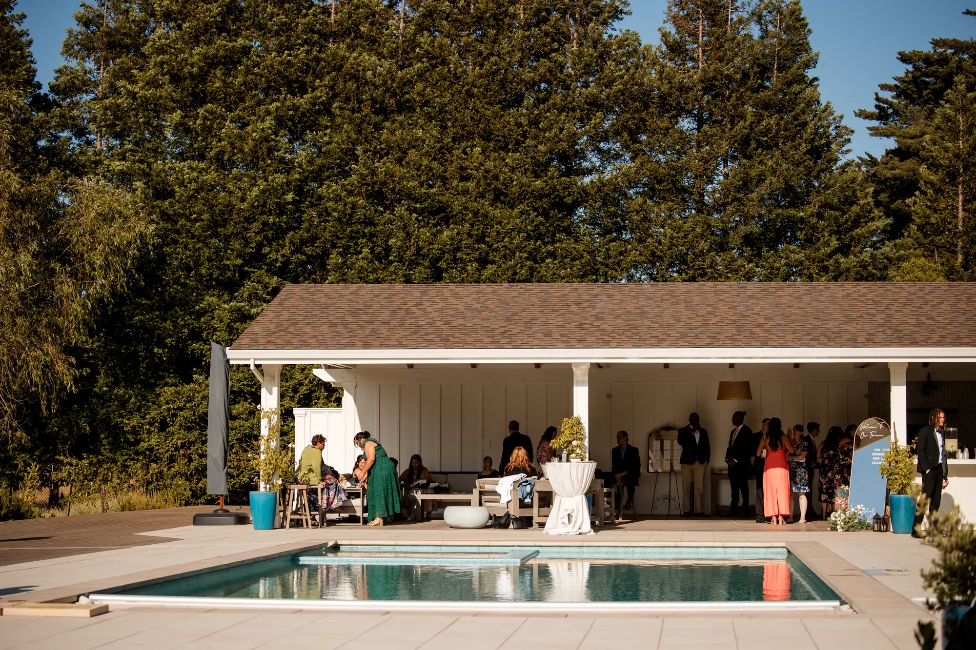 People gathering at an outdoor poolside event near a white pavilion, with trees in the background, and a swimming pool in the foreground.