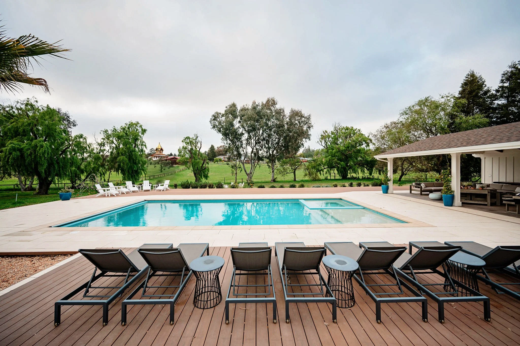 Outdoor swimming pool area with black lounge chairs and small tables on a wooden deck, surrounded by trees and greenery under a cloudy sky.