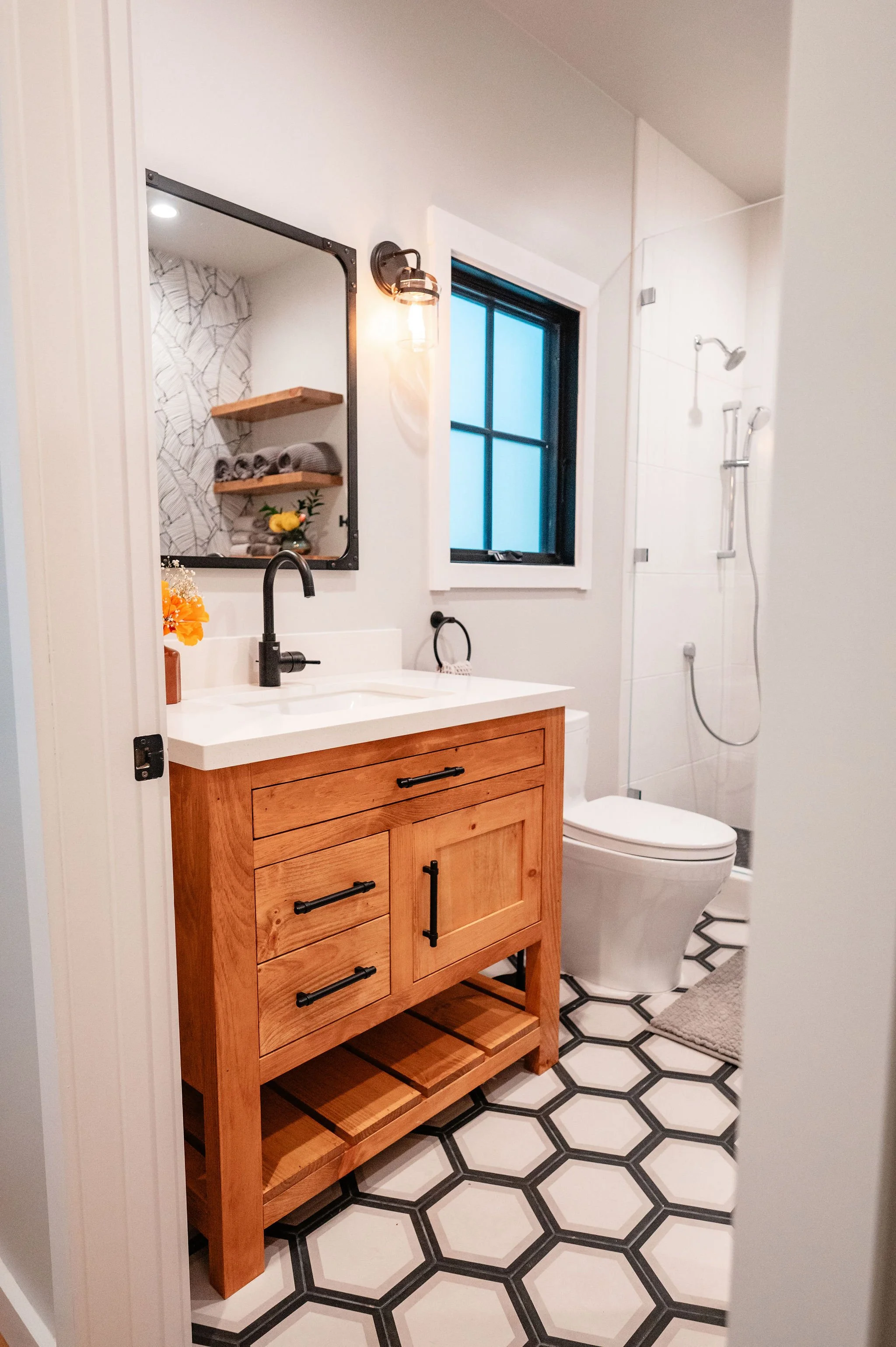 Bathroom with wooden vanity, black faucet, grey towels, mirror, wall sconce, window, toilet, and shower area with glass door.