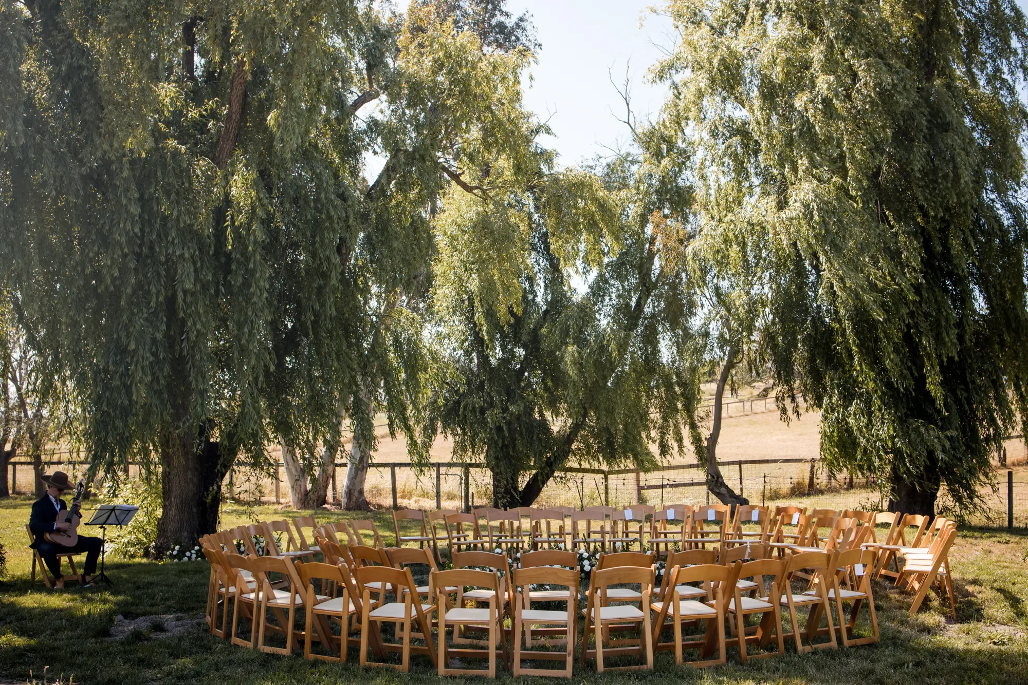 An outdoor wedding ceremony setup with wooden chairs arranged in a circle, decorated with flowers. A musician is sitting on a chair under trees, playing a guitar. The scene is sunny with lush greenery and a field in the background.