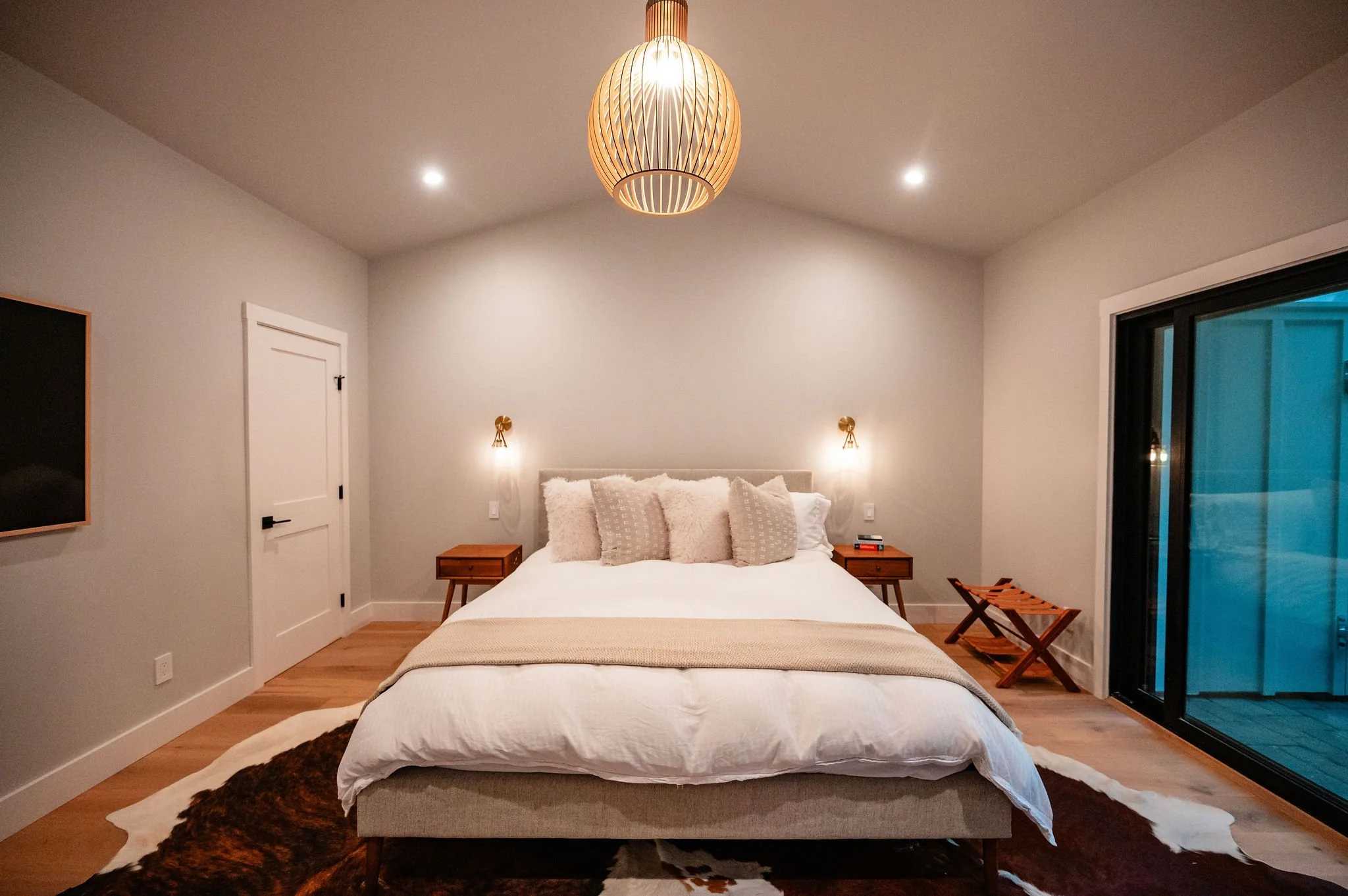 A modern bedroom with a bed having multiple pillows, two wall-mounted lamps, two wooden nightstands, a sliding glass door on the right, and a cowhide rug on the wooden floor.