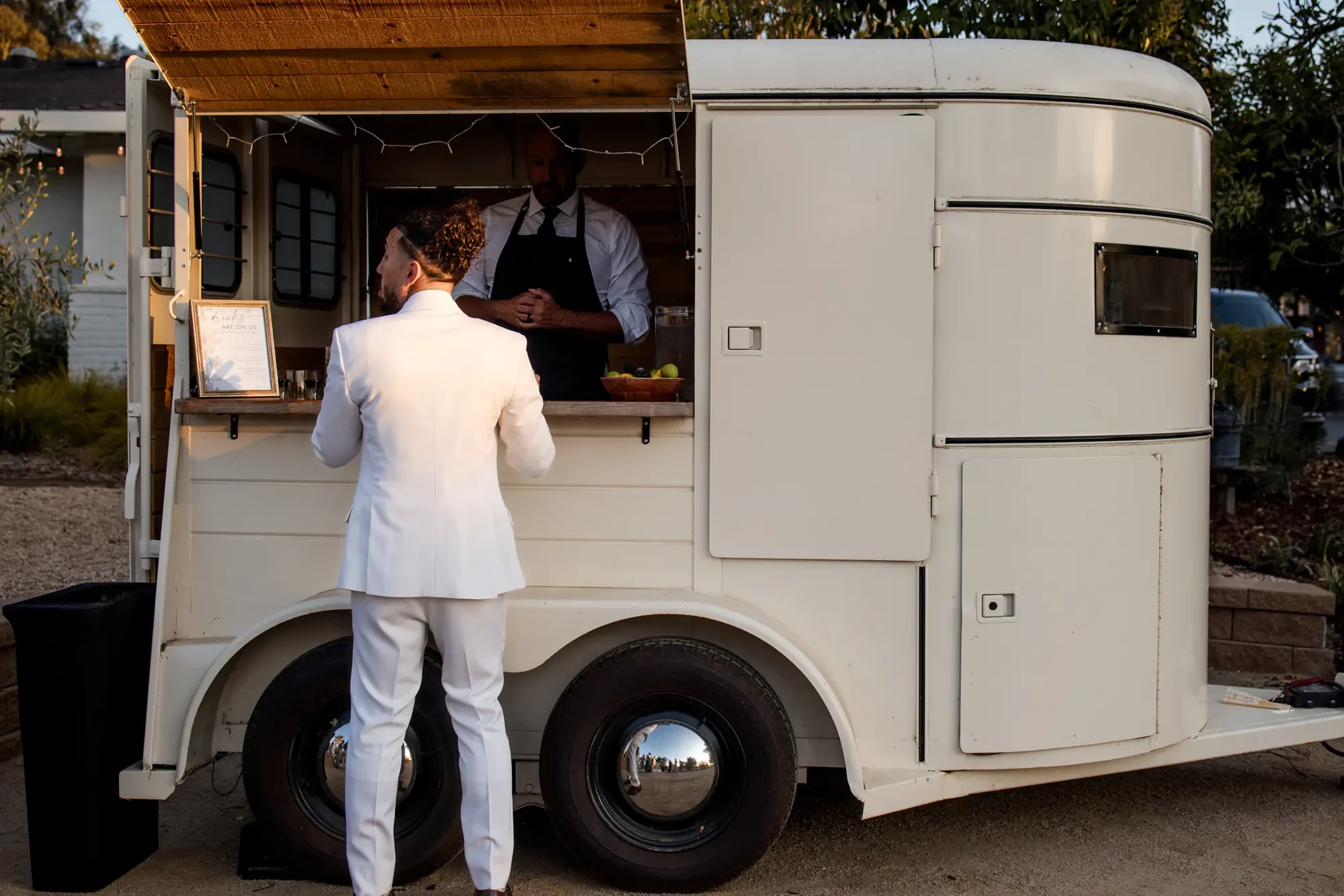 A person in a white suit standing at a food truck window, talking to a man inside who is wearing a black apron, with trees and a house in the background.