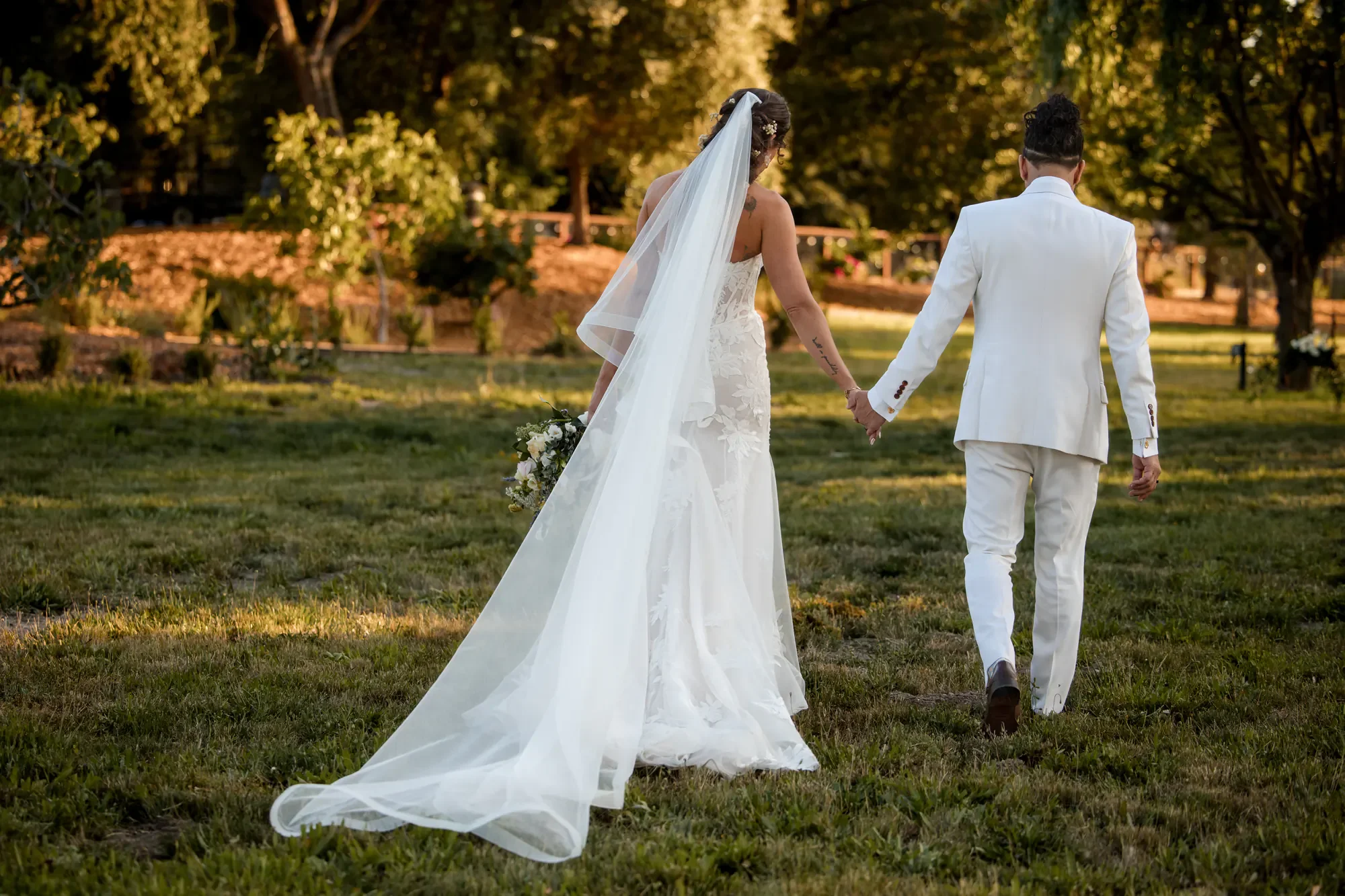 A bride and groom walking hand in hand outdoors in a grassy area, with trees in the background, during sunset.