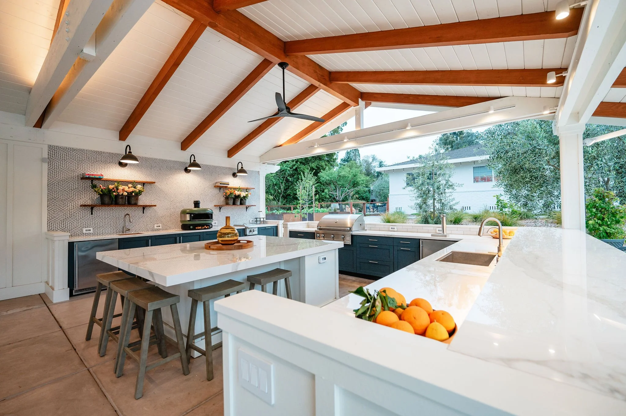 Modern outdoor kitchen with white countertops, black cabinets, and open views of trees and neighboring houses, featuring a ceiling fan, pendant lighting, and fresh oranges on the counter.