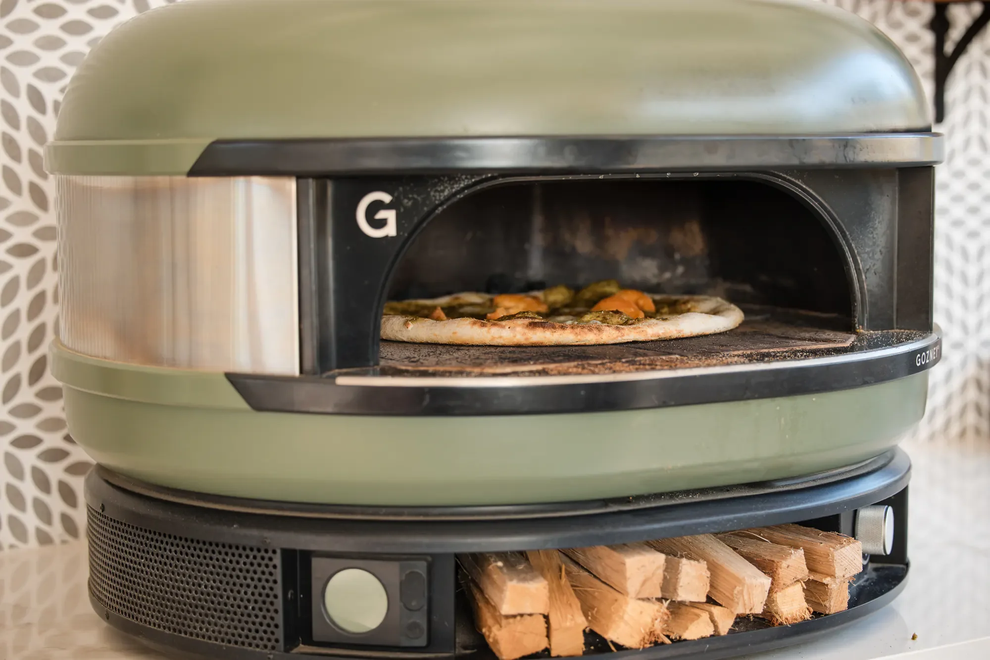 A green pizza oven with a black opening, baking a pizza topped with vegetables. Below the oven, there is a space with chopped wood logs.