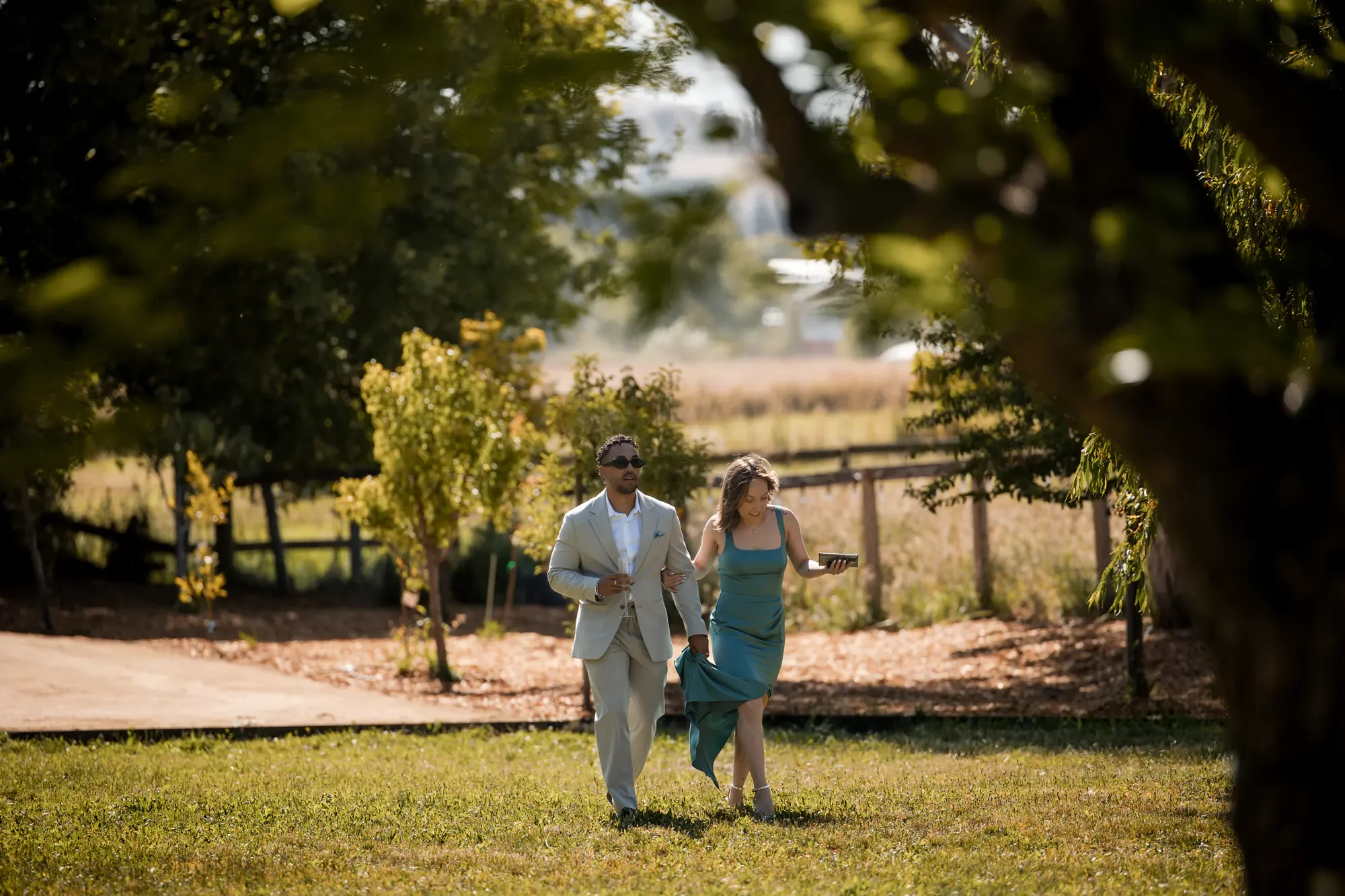 A man in a light-colored suit and sunglasses walks with a woman in a teal dress, who is holding a menu or card, through a grassy area with trees and a wooden fence in the background on a sunny day.