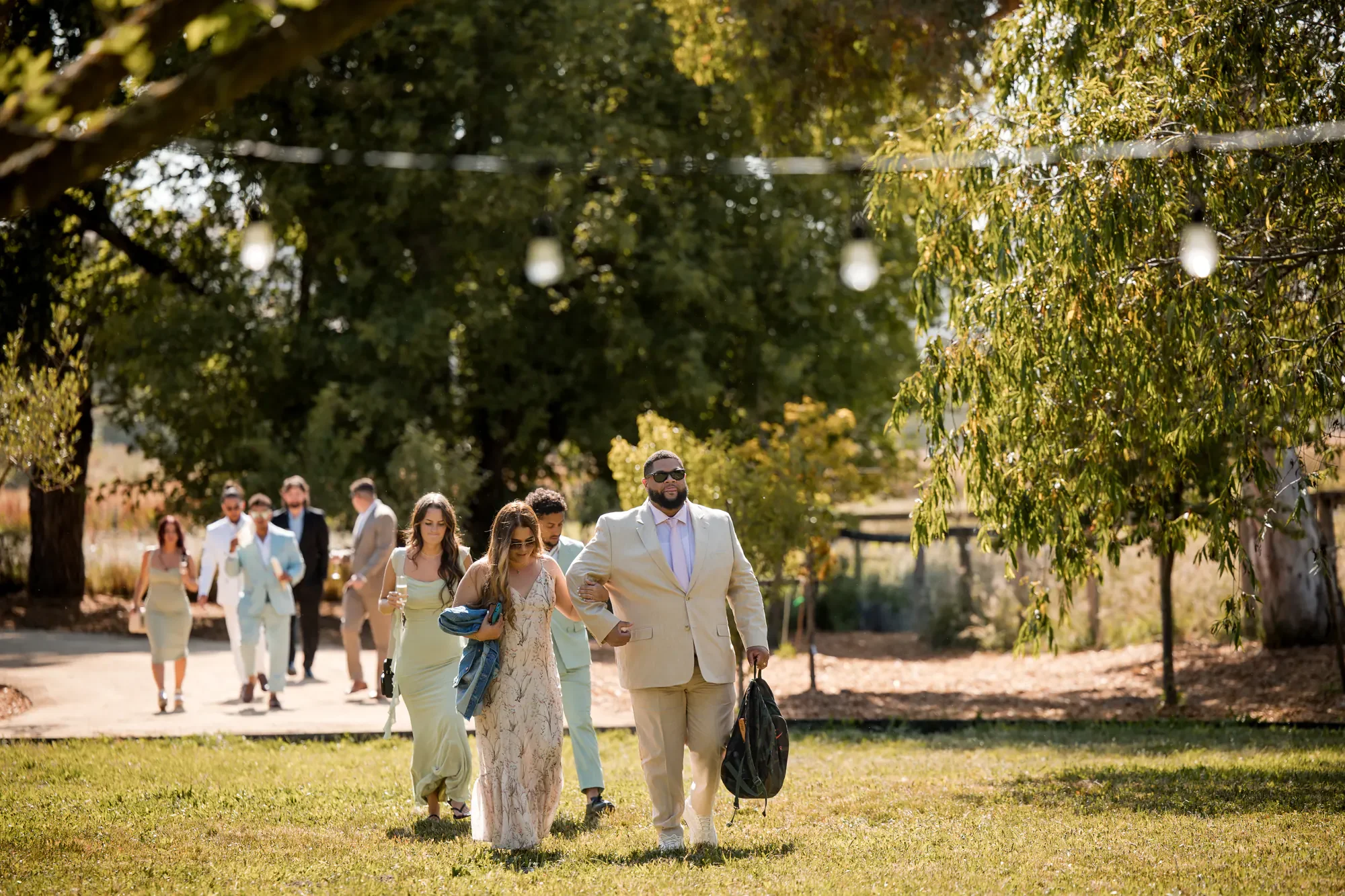 Group of people dressed in formal and semi-formal attire walking outdoors on a sunny day with green trees and grass.