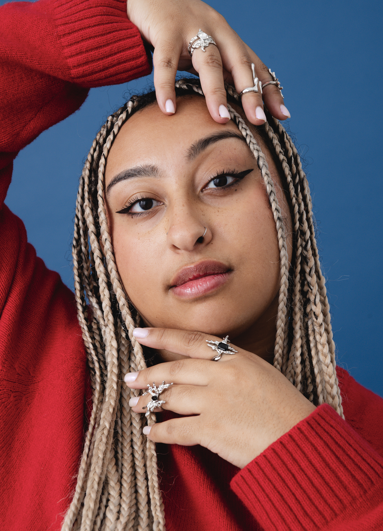 A young woman with braid hair and light makeup, wearing a red sweater, jewelry, and black eyeliner, poses against a blue background with one hand on her face and the other on her forehead.