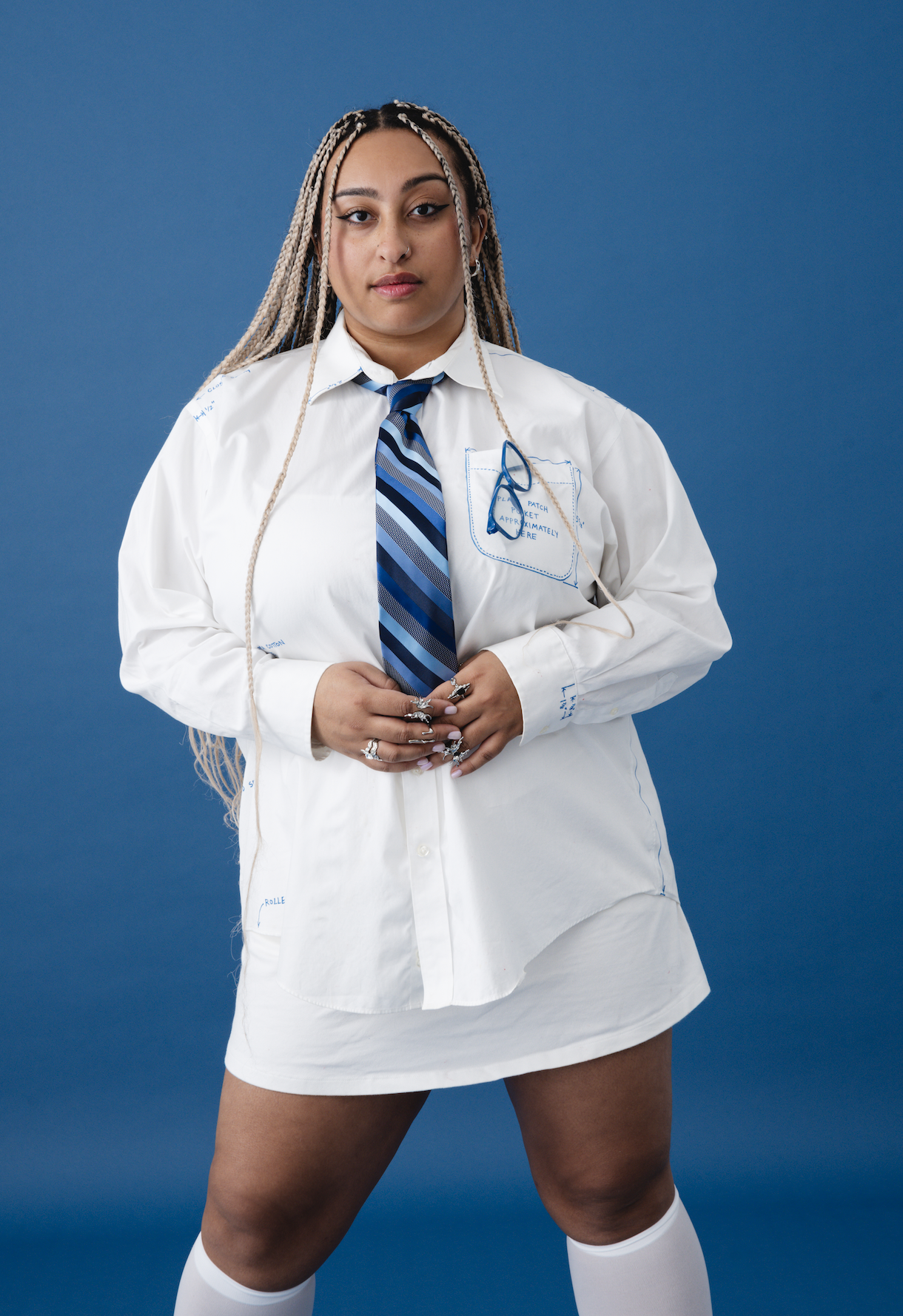 A young woman with long blonde braids wearing a white shirt, blue striped tie, and white knee-high socks standing against a blue background.