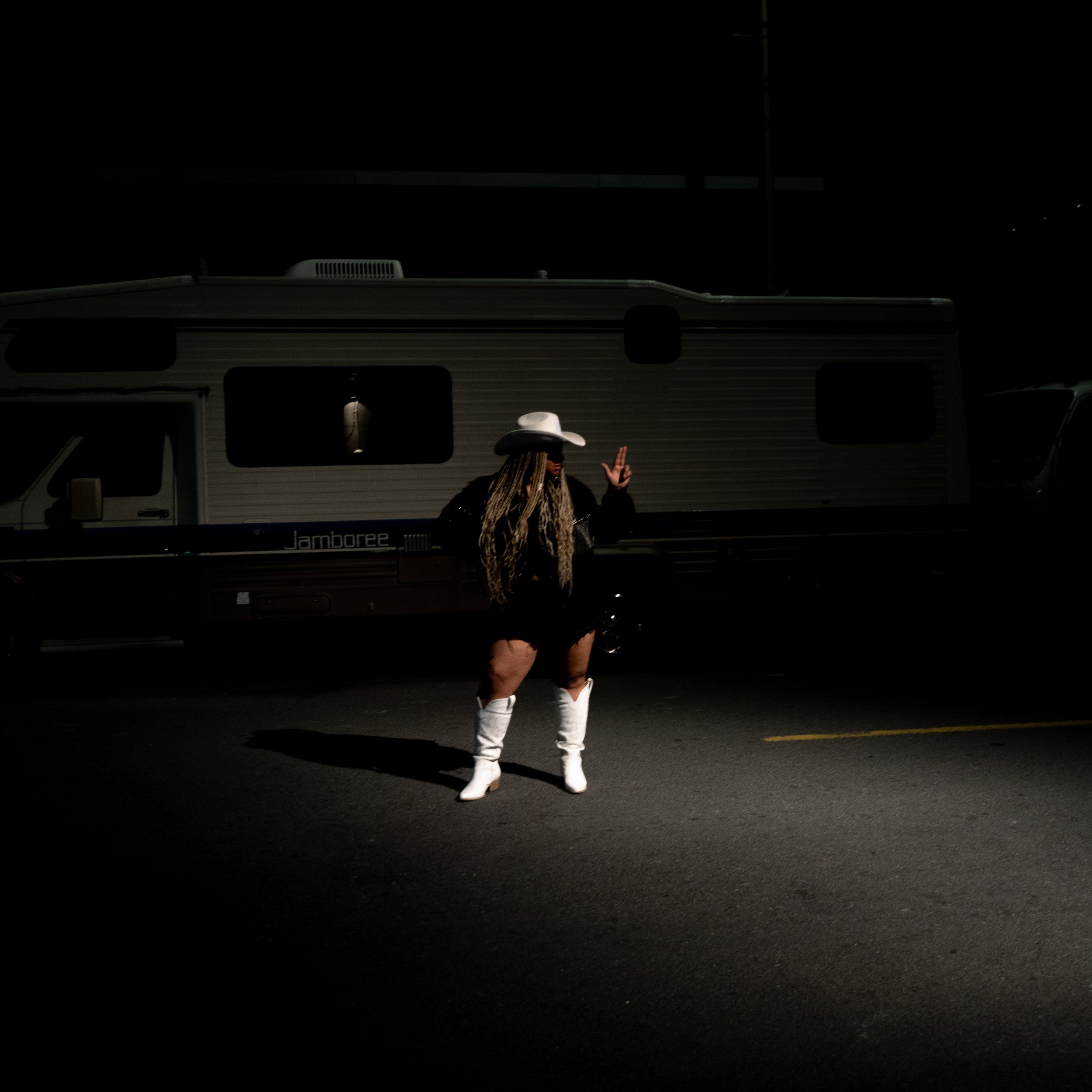 Person standing in front of a recreational vehicle at night, wearing a white cowboy hat, black outfit, white boots, and making a peace sign.