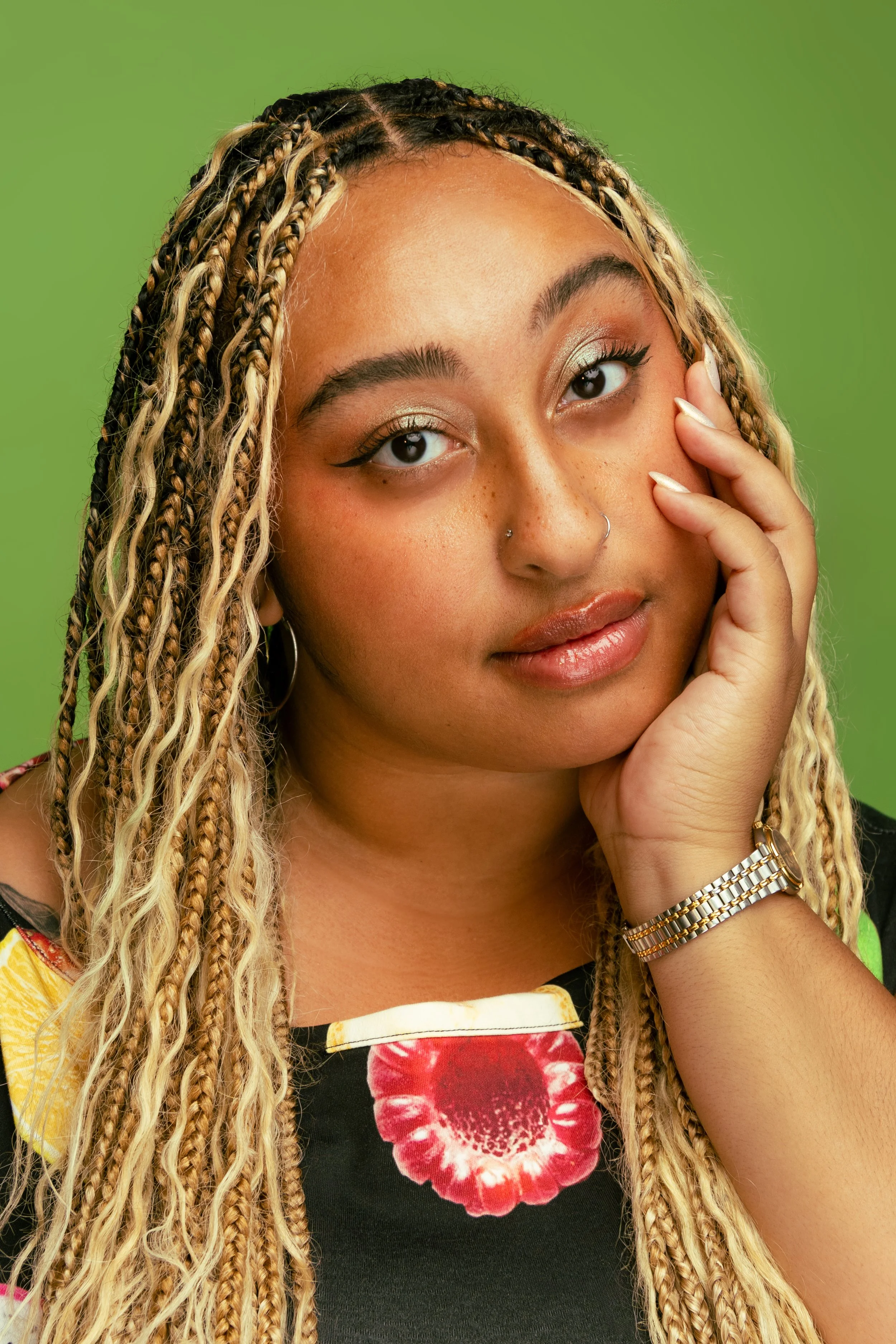 A young woman with long blonde braids, gold eye makeup, light lipstick, and a nose piercing, resting her face on her hand against a green background.