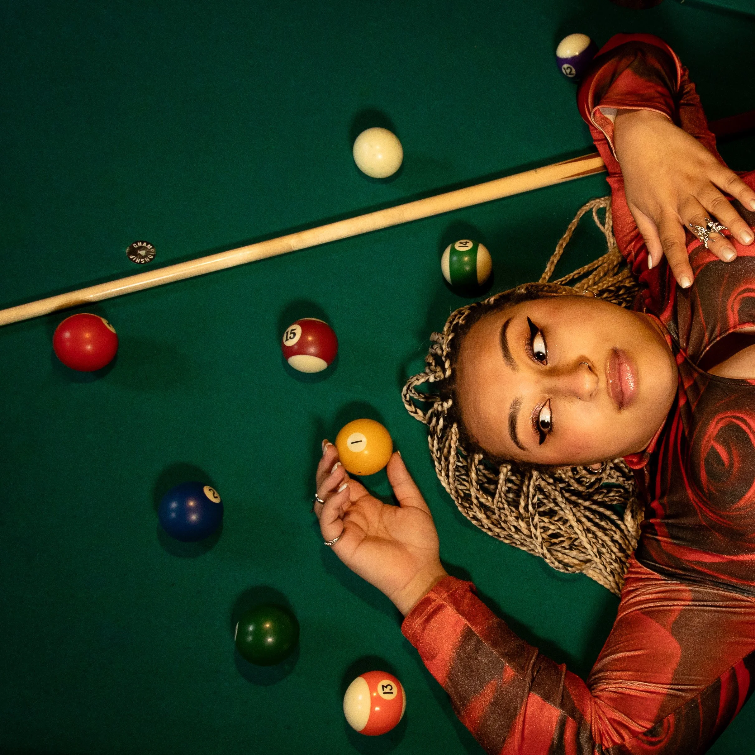 A woman lying on a green pool table surrounded by billiard balls, holding the 1 yellow ball, with a pool cue and a black 8 ball nearby, and wearing a red and black outfit with braided hair.