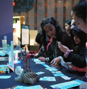 Group of children gathered around a table with various items and informational materials, engaging with a display or exhibit at an indoor event.