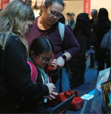 A young girl with glasses and a backpack is using a vintage red rotary phone at an event, with an adult woman looking on. There are other people in the background.