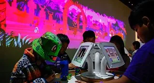 Children using tablets at a tech exhibition with a colorful digital display in the background.