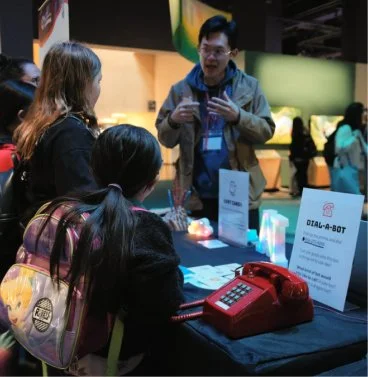 A man demonstrating a product at a booth to two young girls, with a red rotary phone and informational signs on the table.