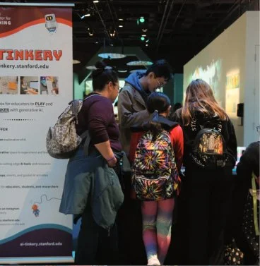 Group of children and adults gathered around interactive exhibit at a science or technology museum, with a poster about tinkering nearby.