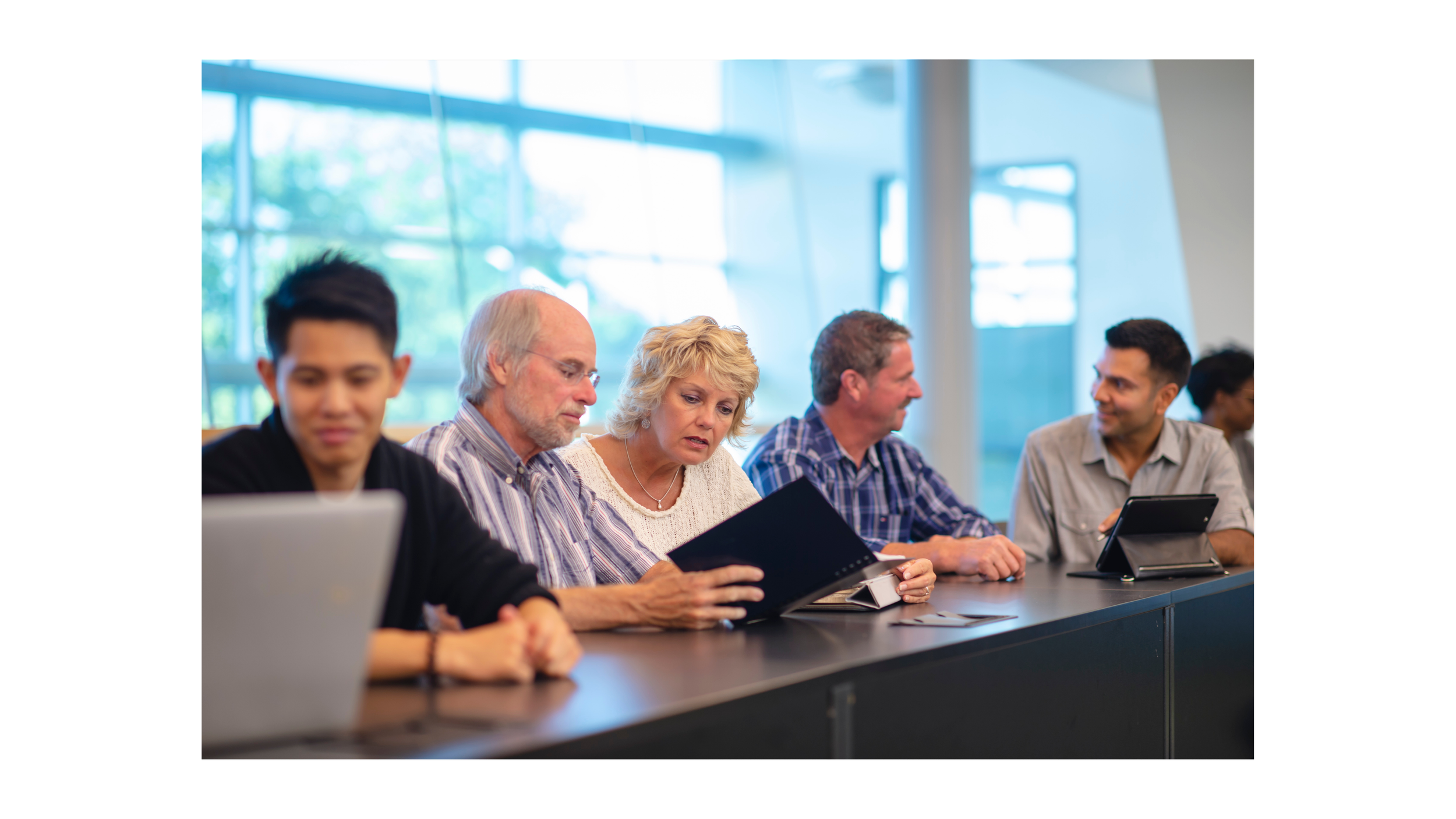 A roomful of professionals seated at a table in a brightly lit room with large windows. Everyone is collaborating and reviewing laptops or tablets.